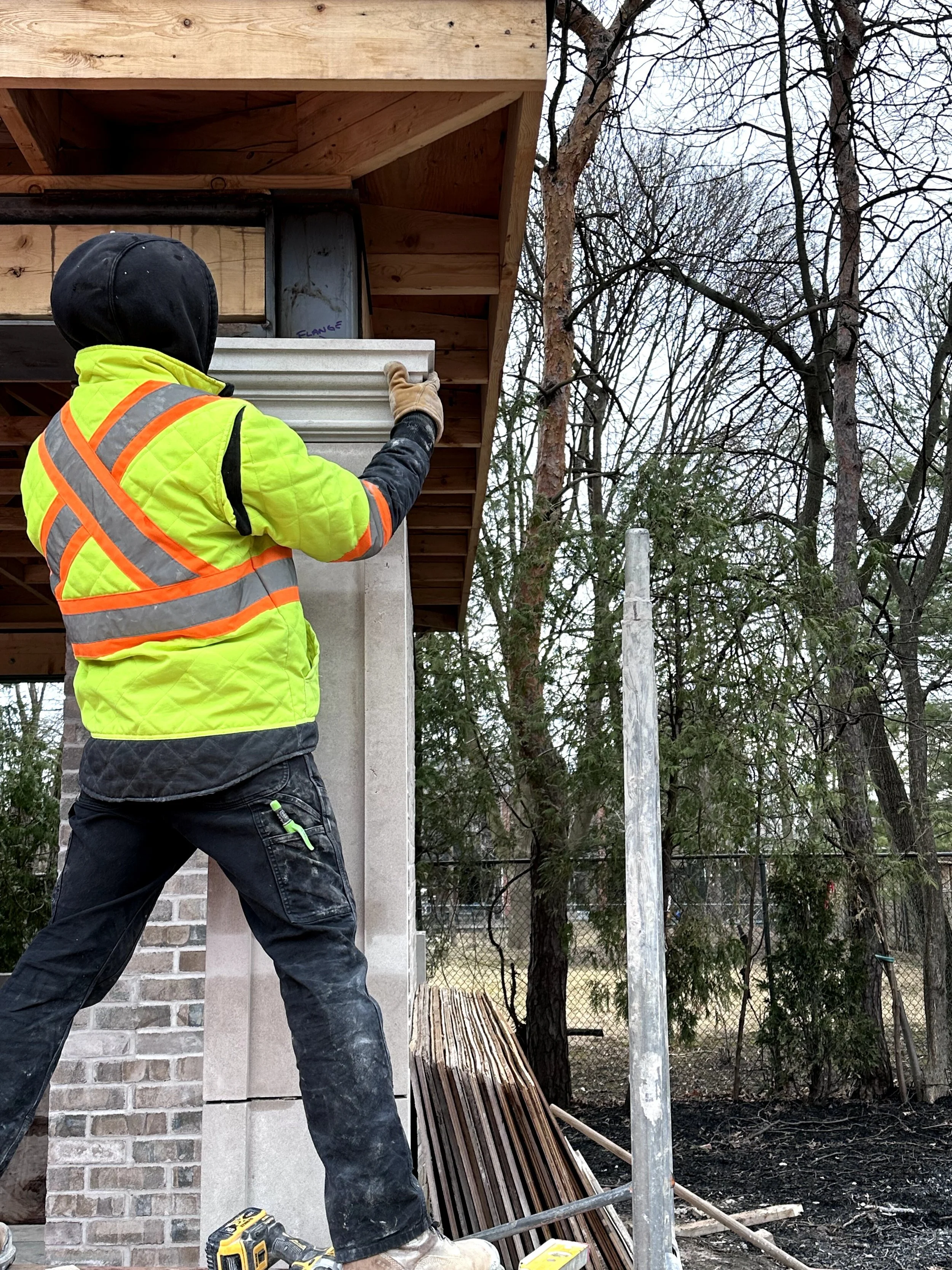 A construction worker in a yellow safety vest and black hoodie is installing a decorative stone wall near a brick building, surrounded by trees and construction materials.