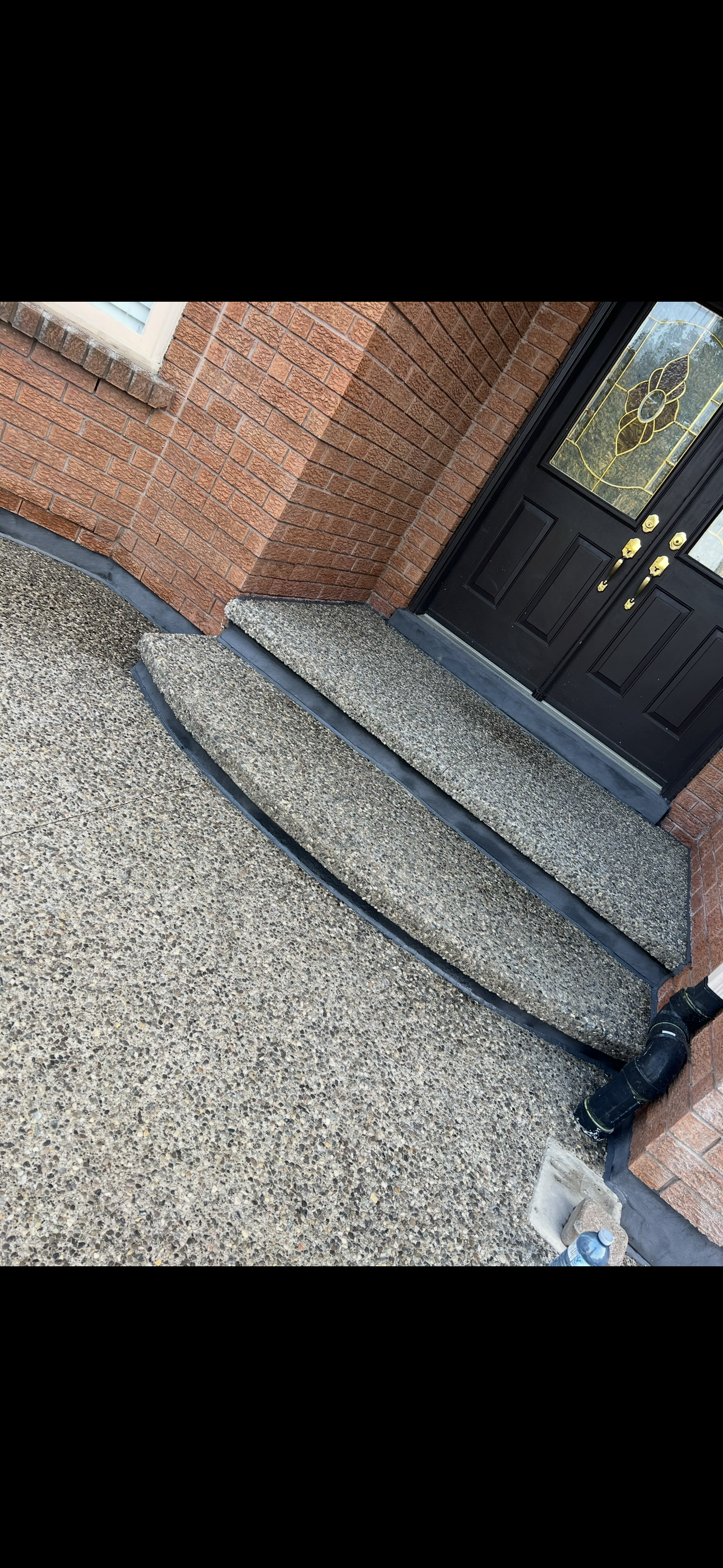 Concrete stairs leading to a black front door with decorative glass panels on a brick house. The ground is textured concrete, and there is a black drainpipe at the bottom right.