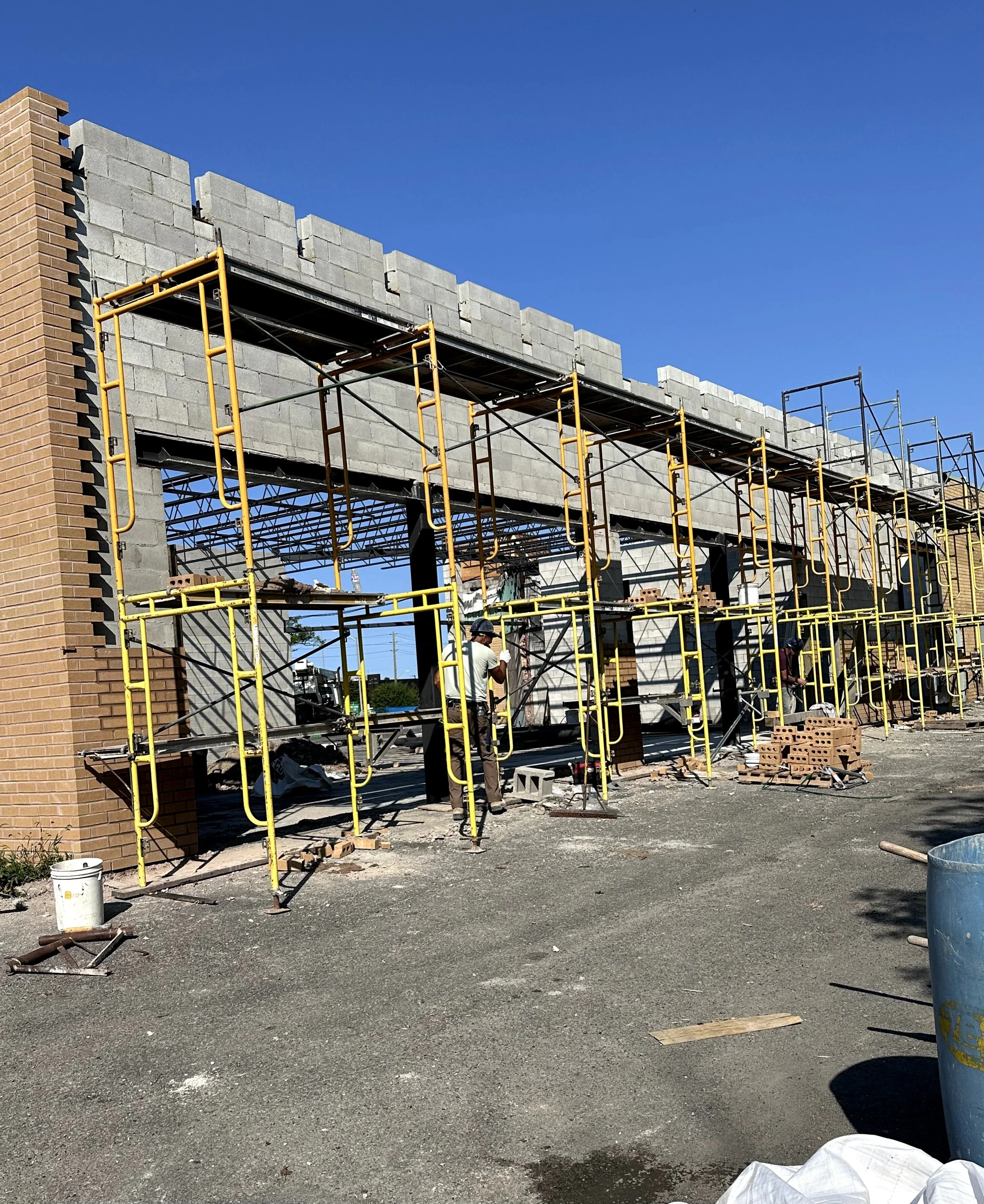 Construction site with scaffolding and partially built brick or concrete structure under a clear blue sky, with a worker in a white shirt and a helmet.