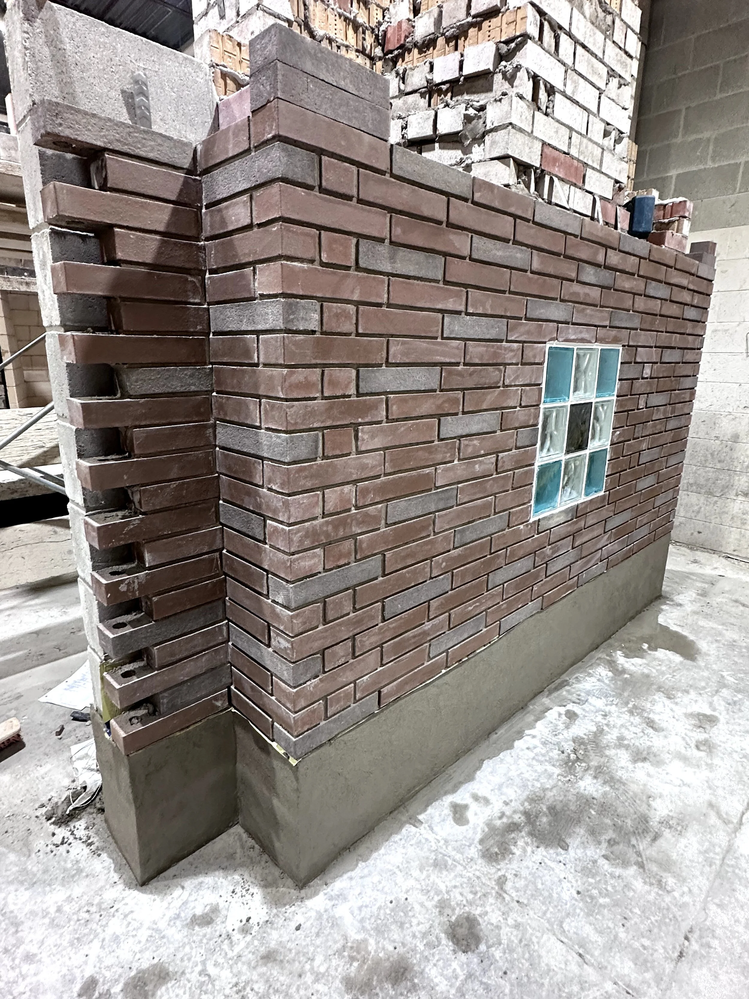 A brick wall under construction with an unfinished corner showing different brick layers and a small window of glass blocks. The ground is concrete with some snow or dust.