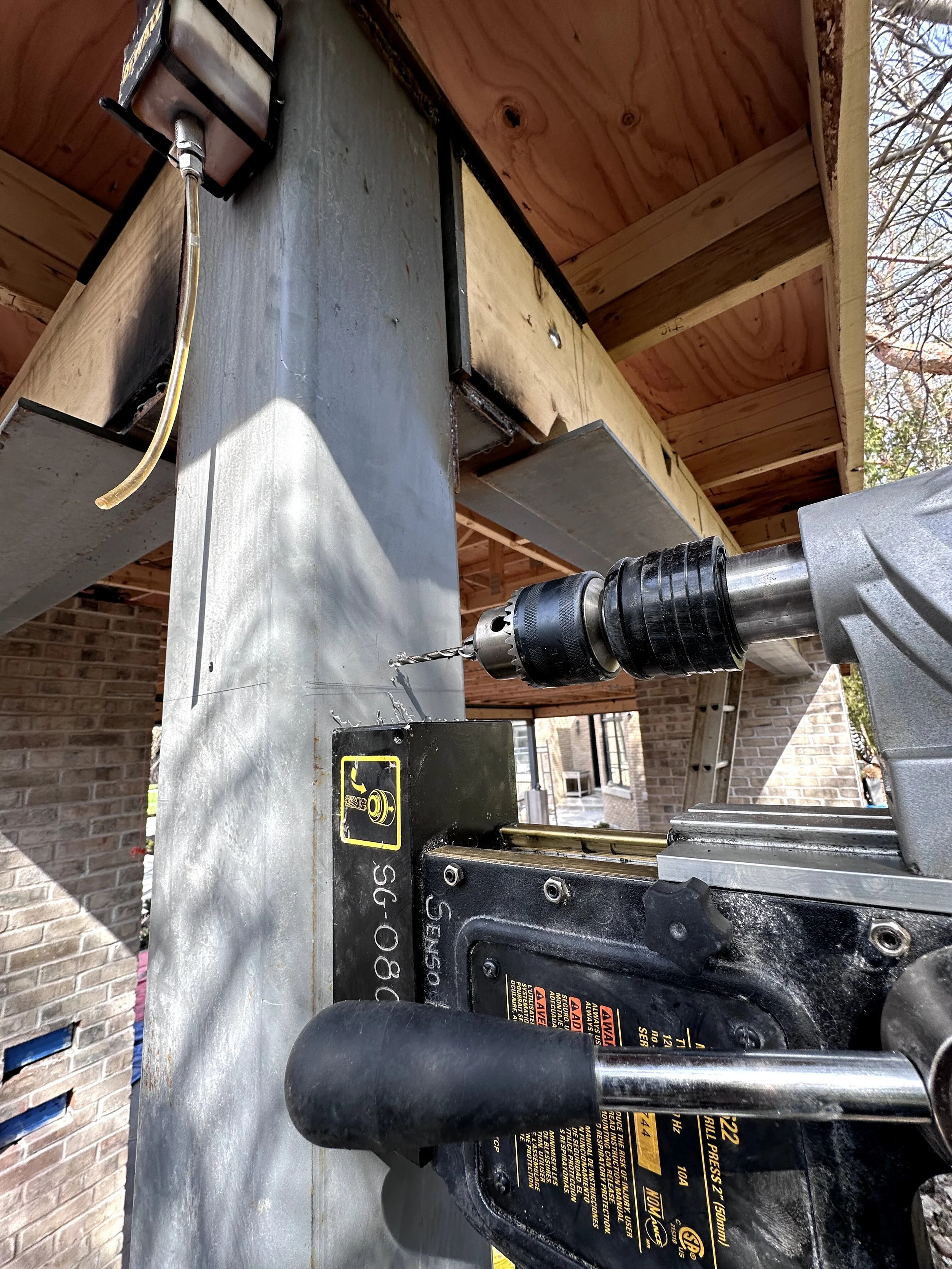Close-up of a power drill mounted on a workbench with safety warnings, working on a metal surface. The background shows a brick wall and part of a wooden roof structure.
