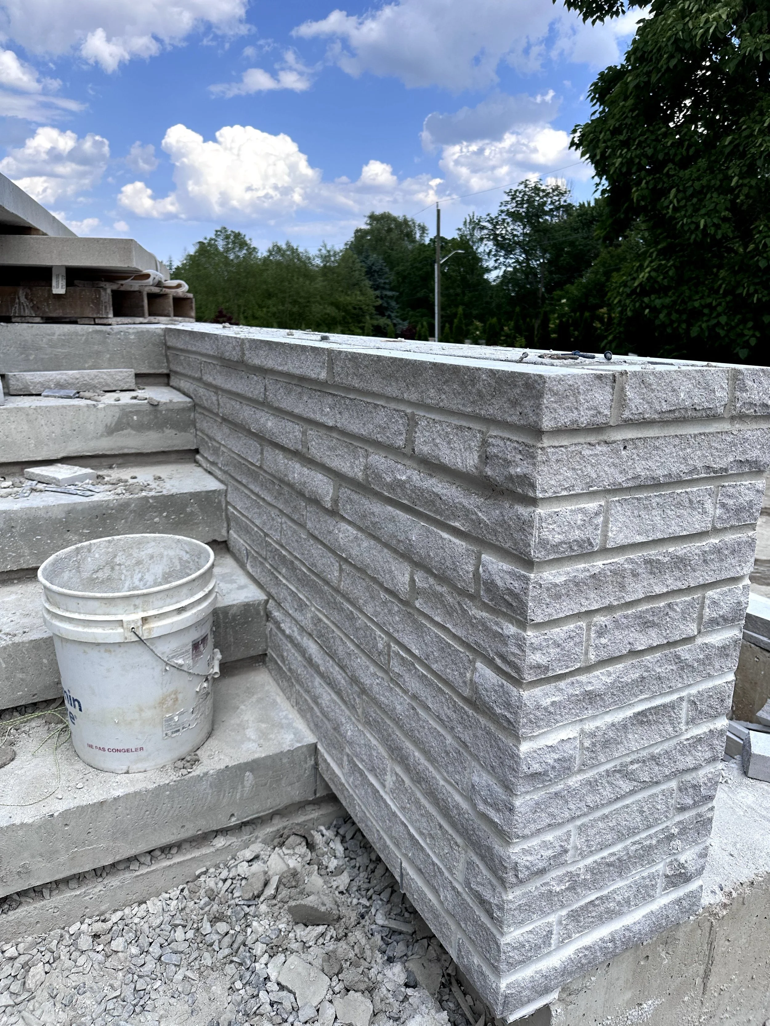 A construction site with a newly built gray brick wall, a white bucket, and scattered debris. In the background, there are green trees, a utility pole, and a partly cloudy sky.