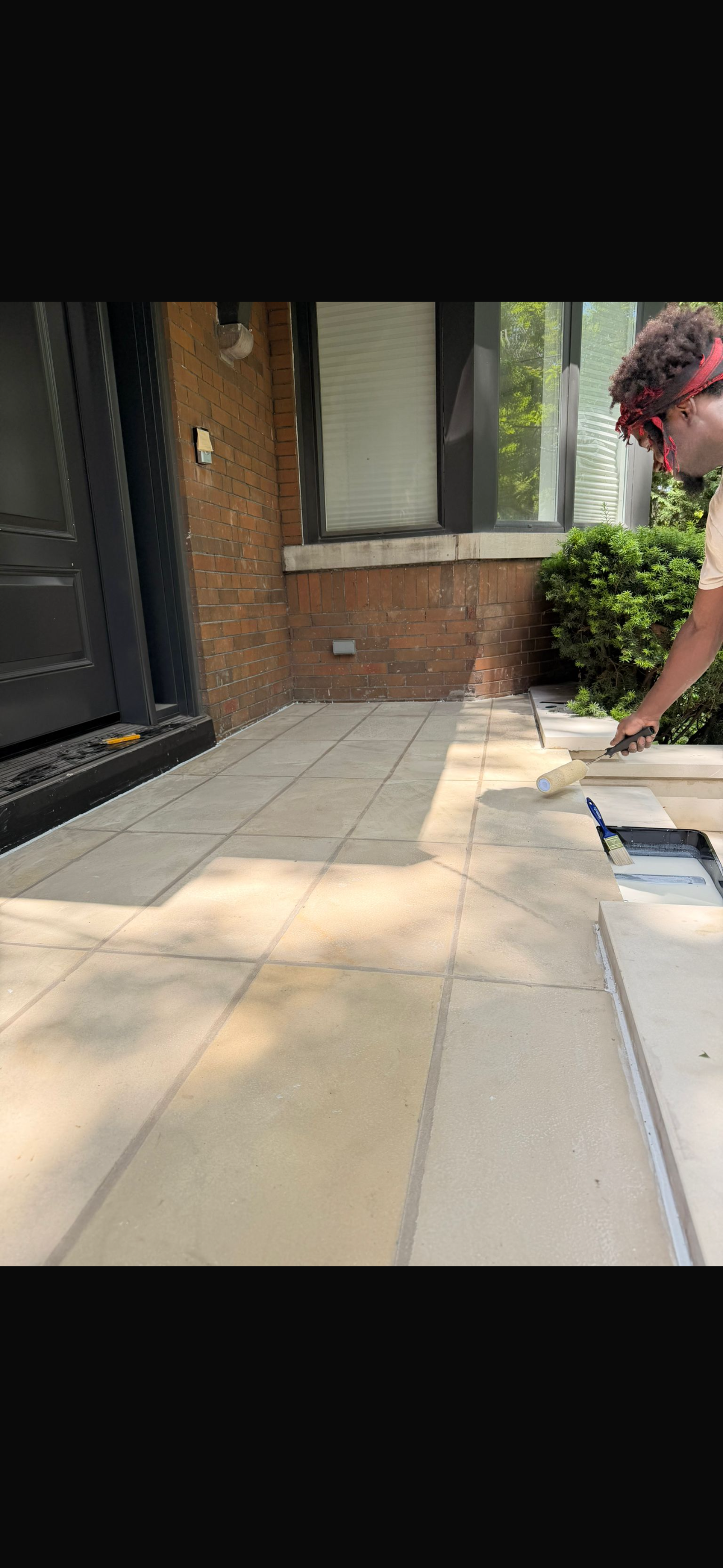 A person painting a white porch step using a paint roller, on a porch with beige tiles, brick wall, and a black door. There's a window and some greenery nearby.
