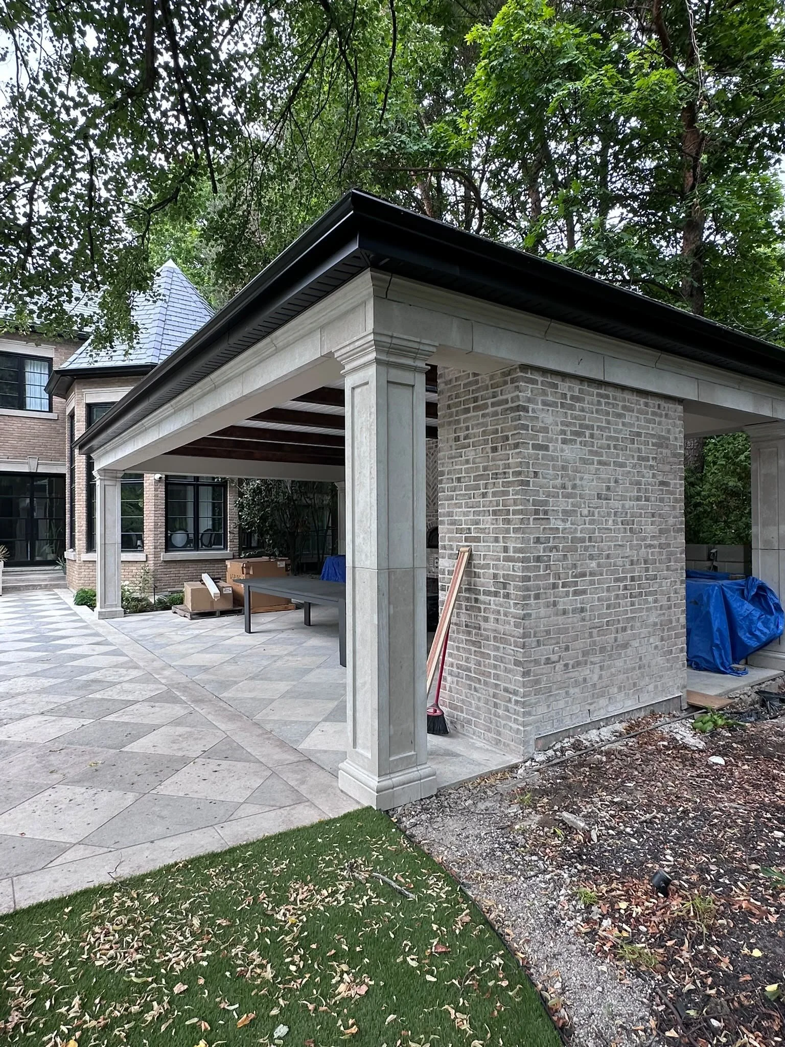A backyard patio area with a stone tiled floor and a brick structure with columns, a roof, and some construction materials and tools around. There are trees and a neighboring building in the background.