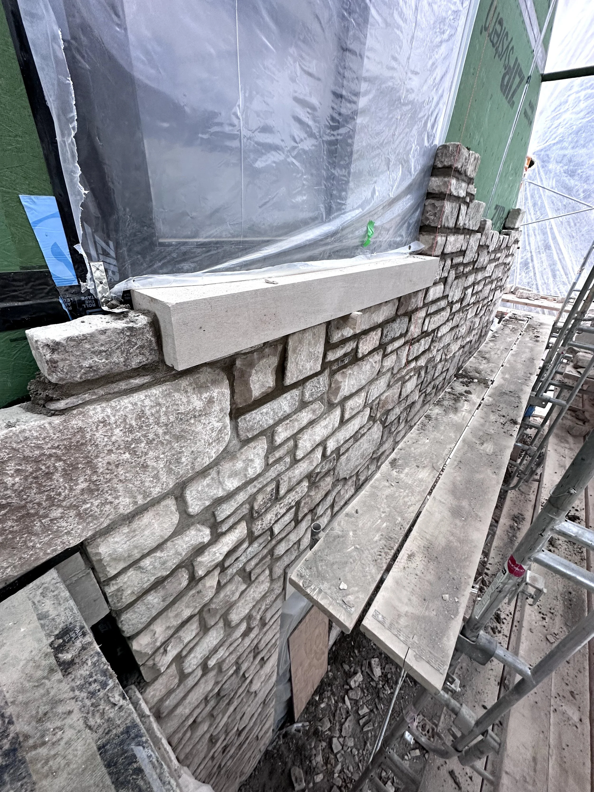 A stone wall under construction with a stone ledge on top, scaffolding on the right side, and a green house wrap on the background wall.