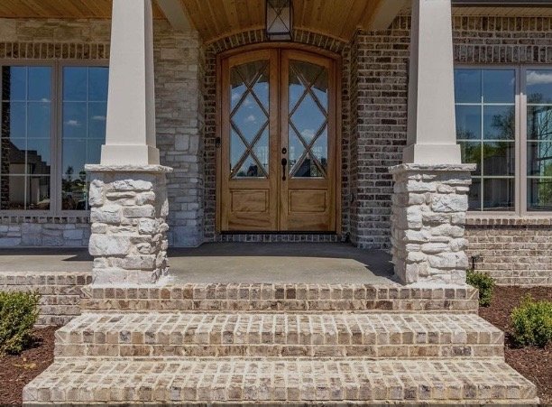 Front entrance of a house with brick steps leading to a wooden double door, flanked by brick and stone columns, with windows on each side.