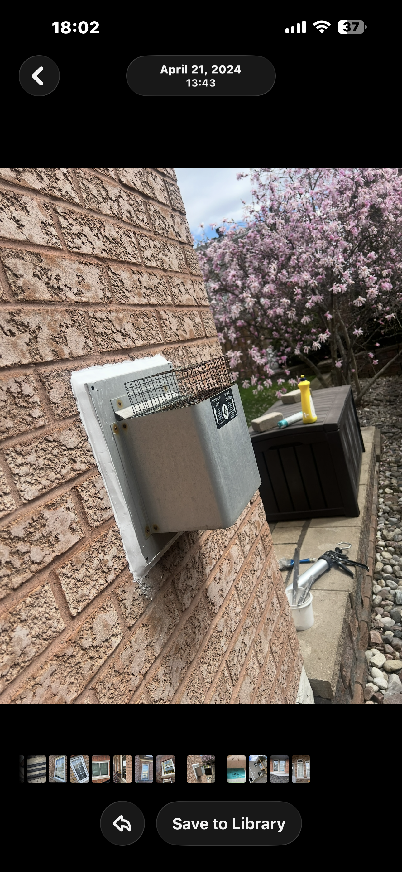 A close-up of a brick wall with an electrical box mounted on it. In the background, there is a garden with a flowering tree with pink blossoms, a black storage container, and gardening tools and supplies on the ground.