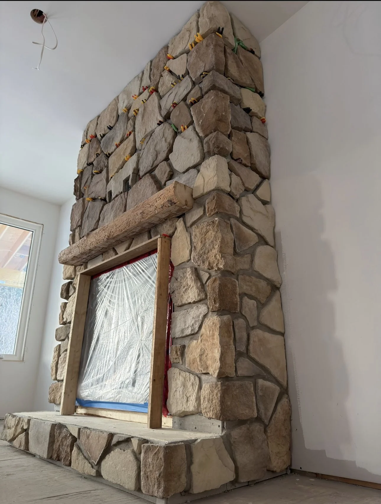 A partially constructed stone fireplace with a wooden mantel and a plastic-covered opening, inside a room with white walls and a window.