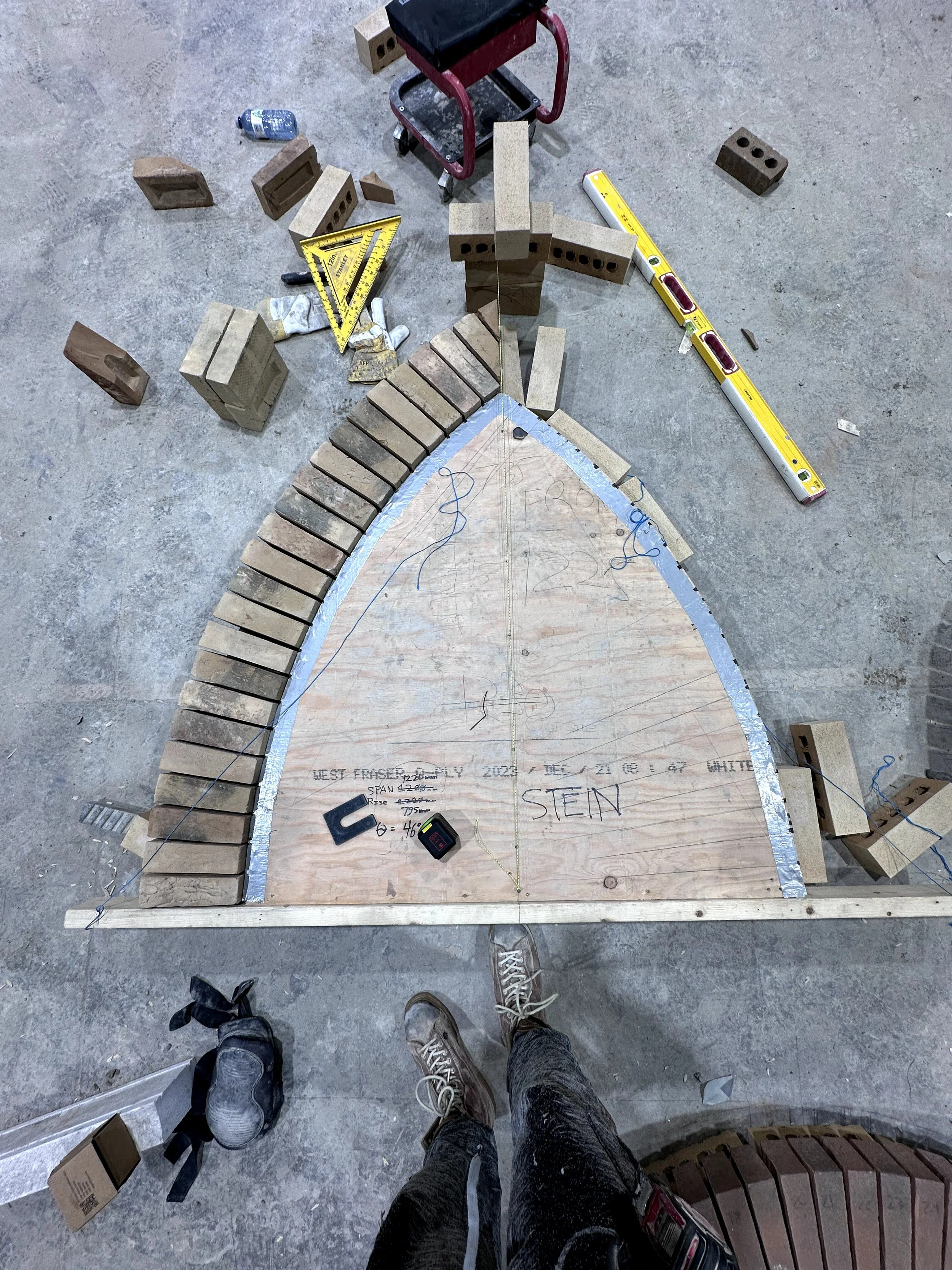 A construction site with a wooden template forming a curved shape, surrounded by bricks, tools, and equipment. Two people wearing sneakers are standing on concrete floor, taking a photo of their work.