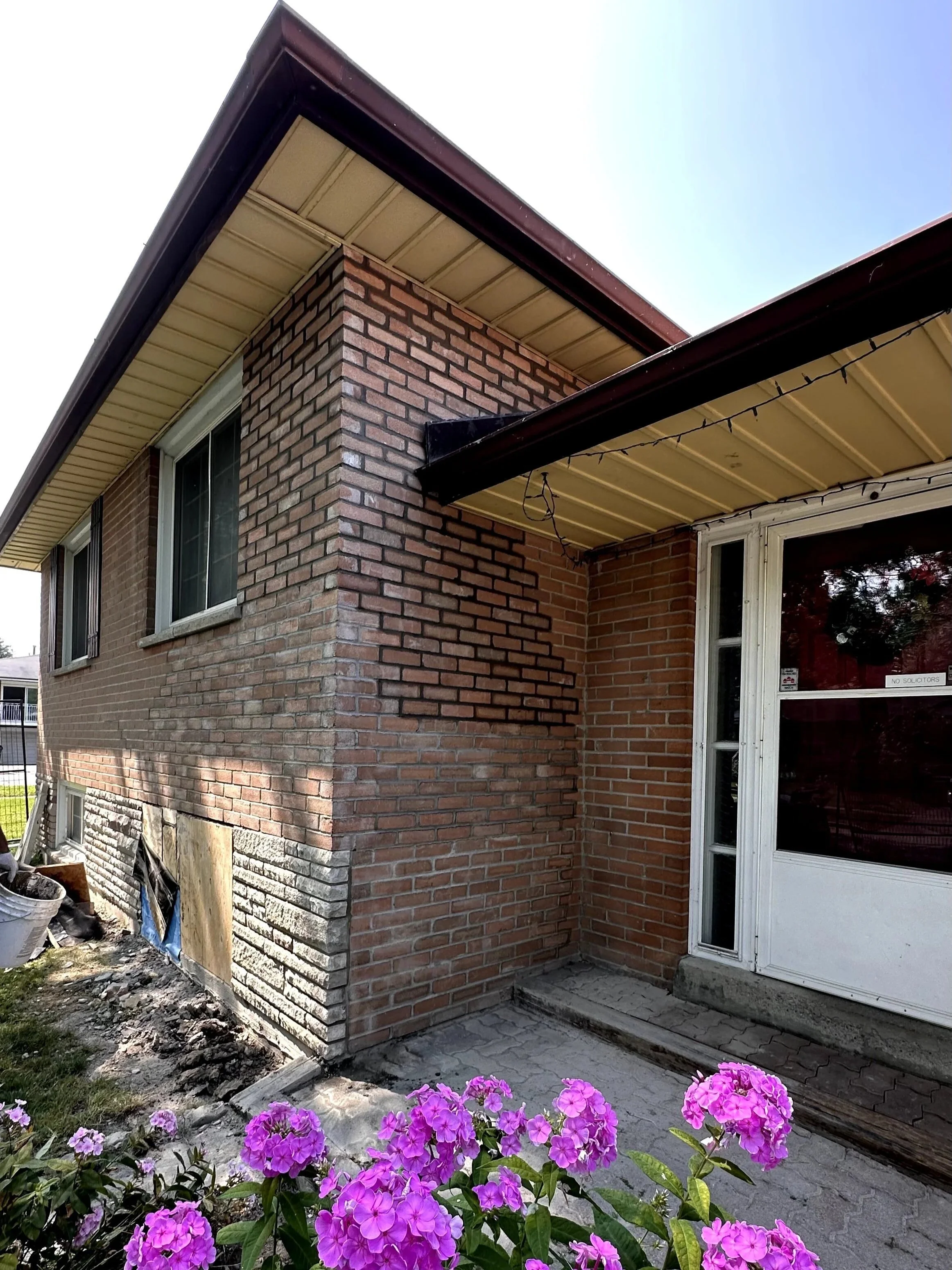 The image shows the corner of a brick house with a window, part of a screened door, and a flower bed with purple flowers in the foreground. The house has a brown roof with gutters, and there are construction materials and debris around the base of the house.