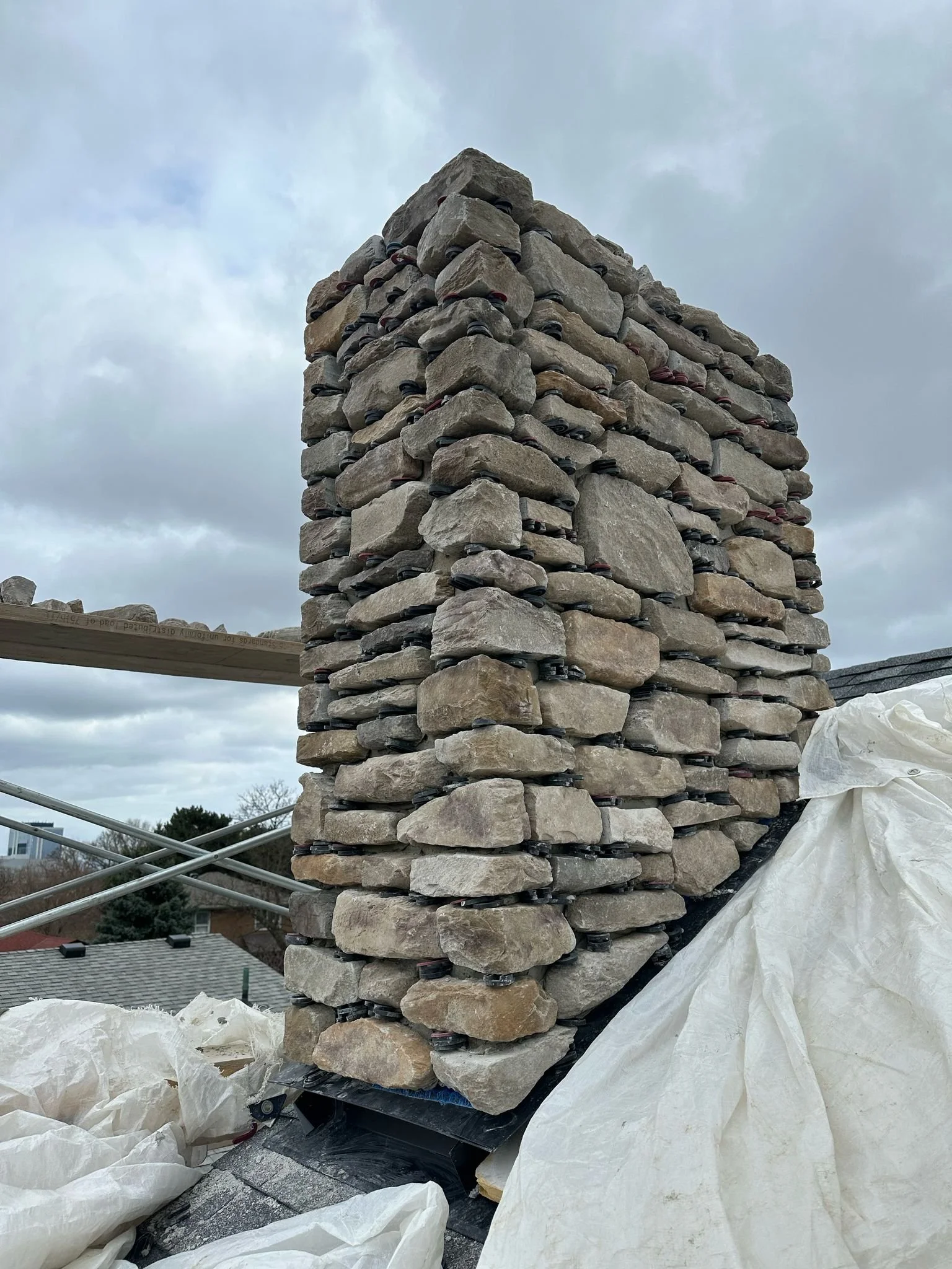 Close-up of a brick chimney under construction or repair with a pile of white construction material, possibly tarp or insulation, around its base. Overcast sky in the background.