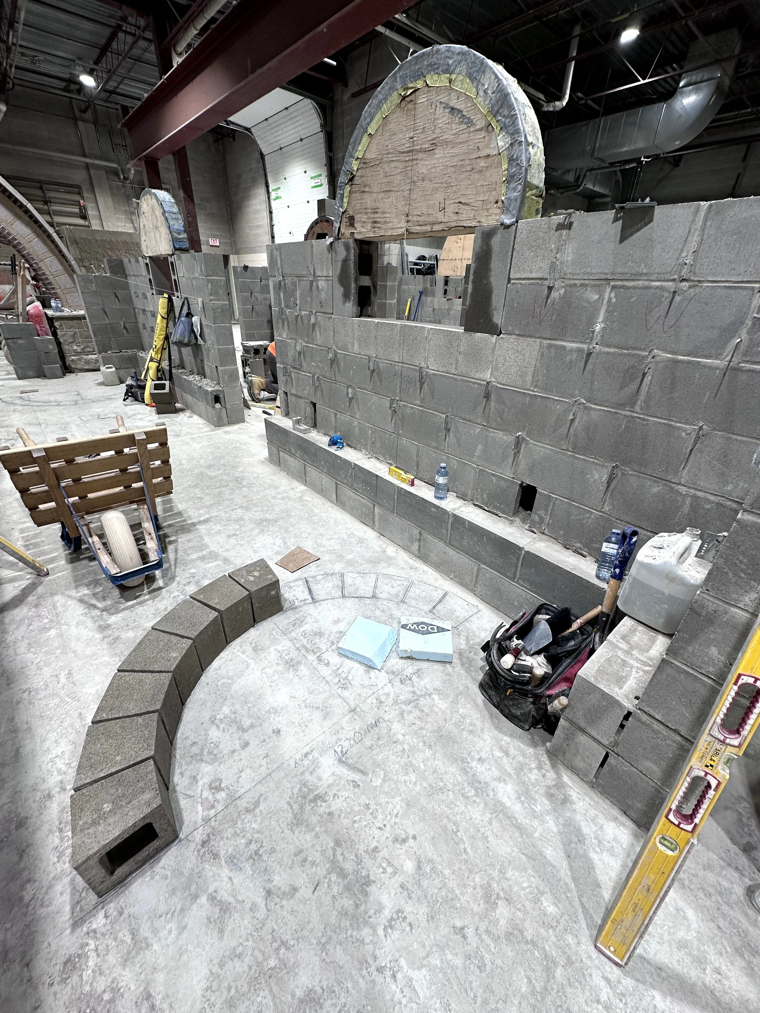 Indoor construction site with cinder block walls, a partially built circular structure, construction tools and materials, and a large yellow level tool leaning against the wall.