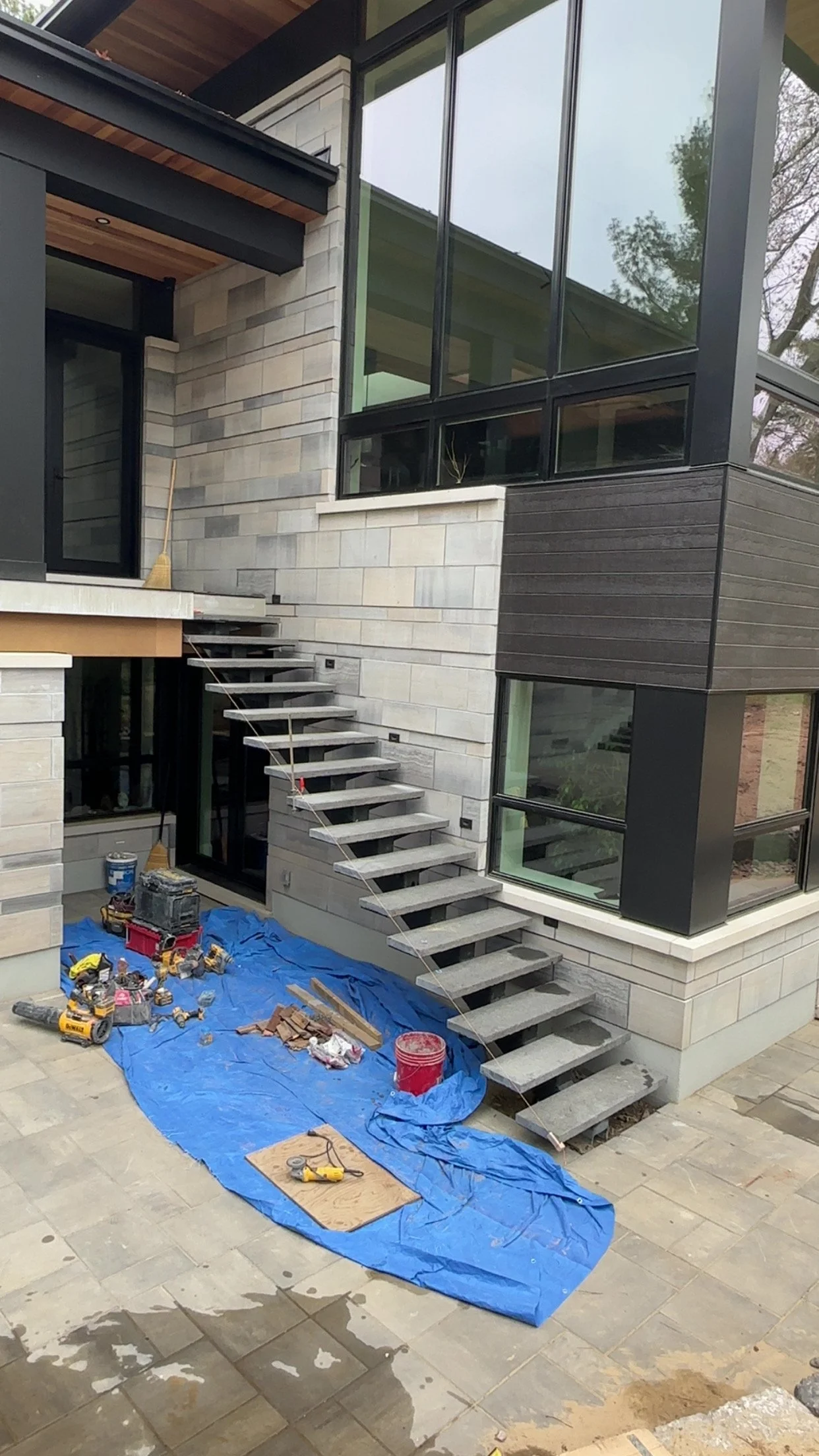 Under construction modern house with large glass windows and stairs leading to upper level, construction tools and materials on blue tarp and tiled ground.