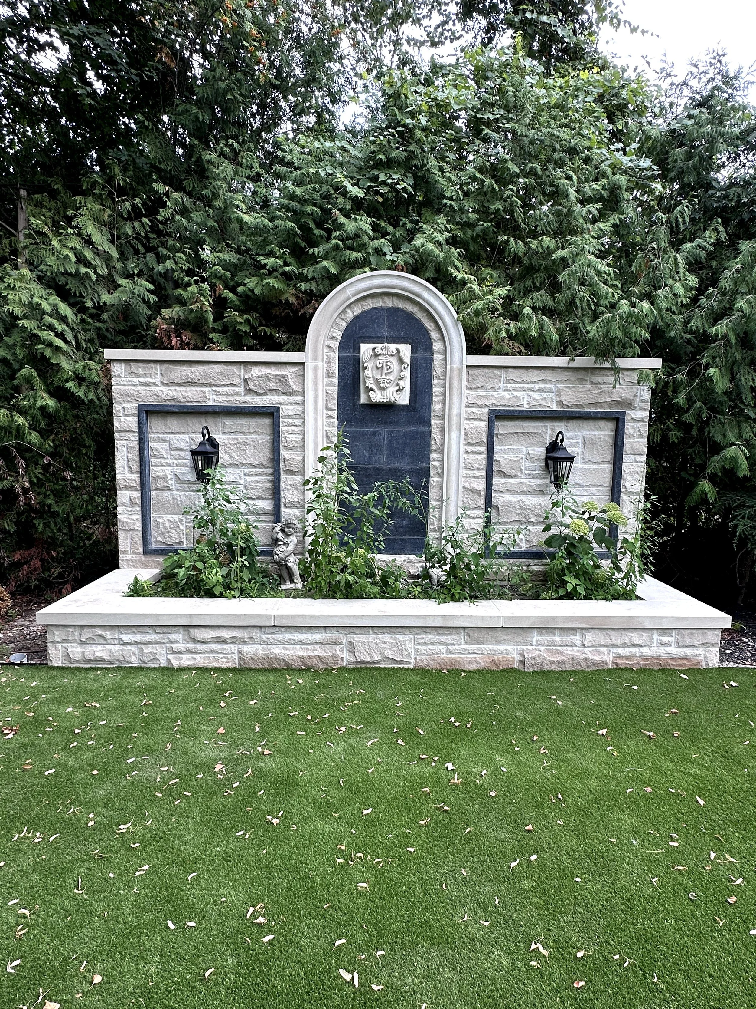 A decorative stone garden feature with a fountain and black lanterns, surrounded by greenery and situated on a manicured lawn.