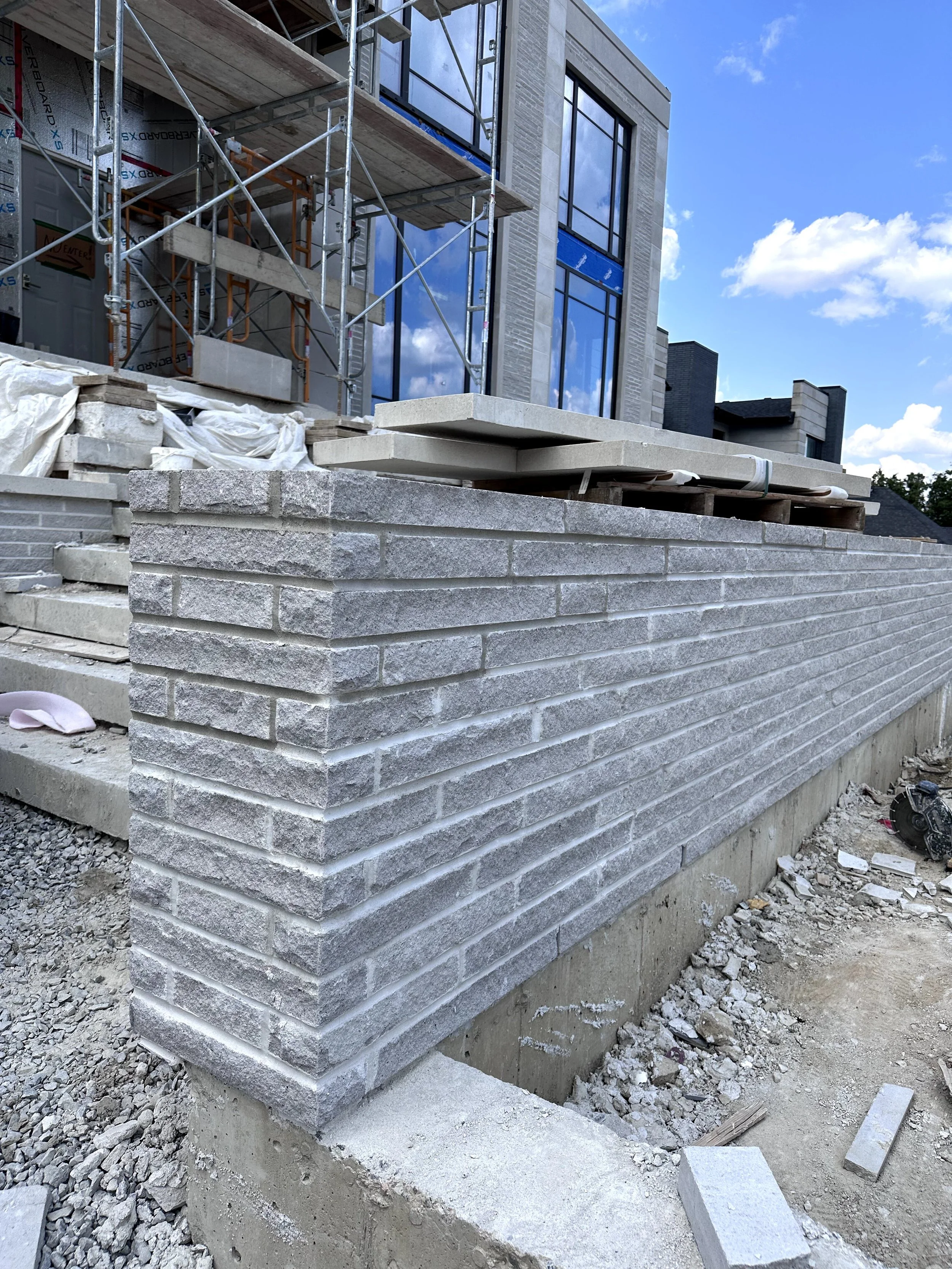 Construction site featuring a brick wall foundation, with stacked bricks and slabs, scaffolding, and a building with large glass windows under construction against a blue sky with clouds.