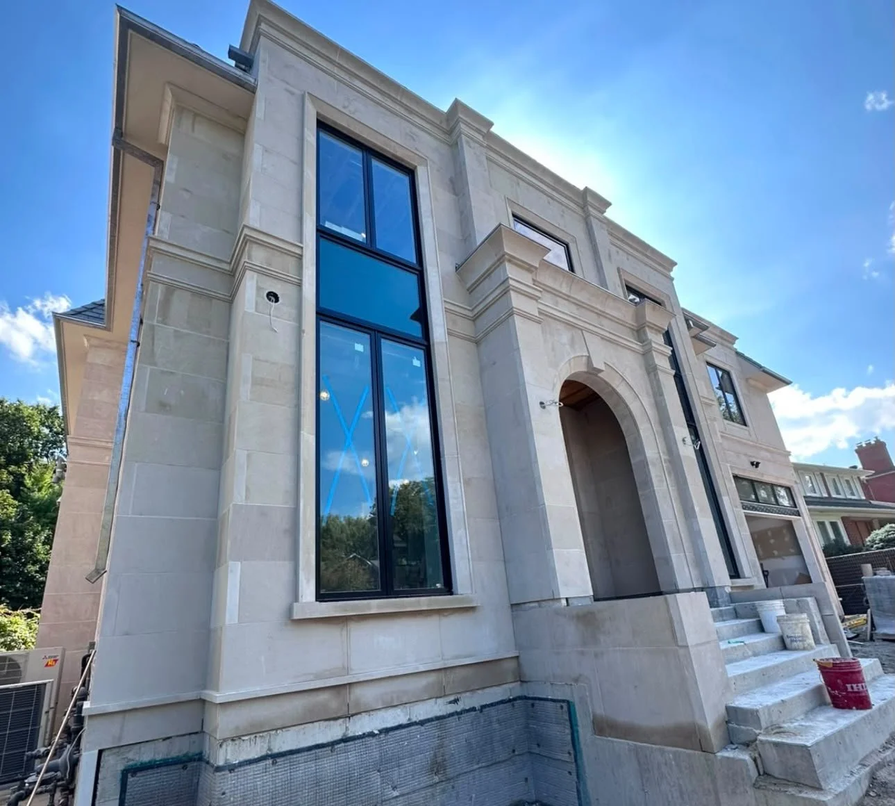 A large unfinished modern house with beige stone exterior walls, tall windows with black frames, and a prominent archway entrance. Construction materials and tools are visible at the front steps, and the sky is clear and blue.