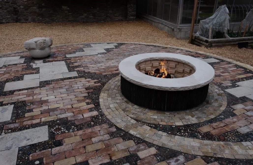 A backyard patio with a circular firepit surrounded by brick and stone paving, and a decorative stone sculpture in the shape of an animal head on the left. There is a gravel area and a greenhouse or garden area in the background.