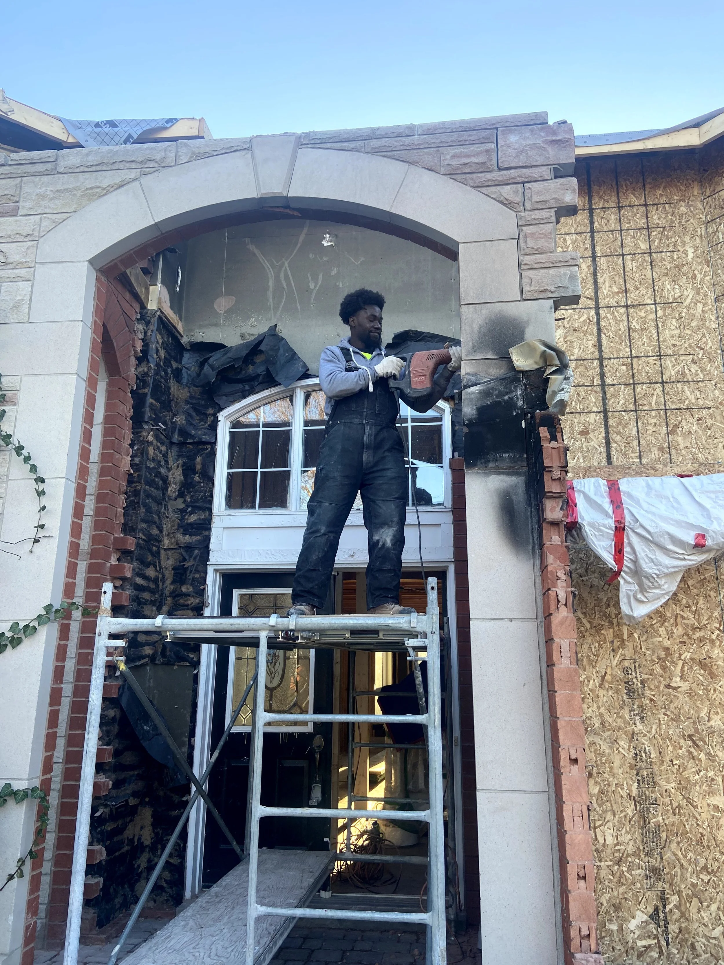 Man working on the construction of a house or building, standing on a scaffold, using a power tool to work on the exterior wall near a window.