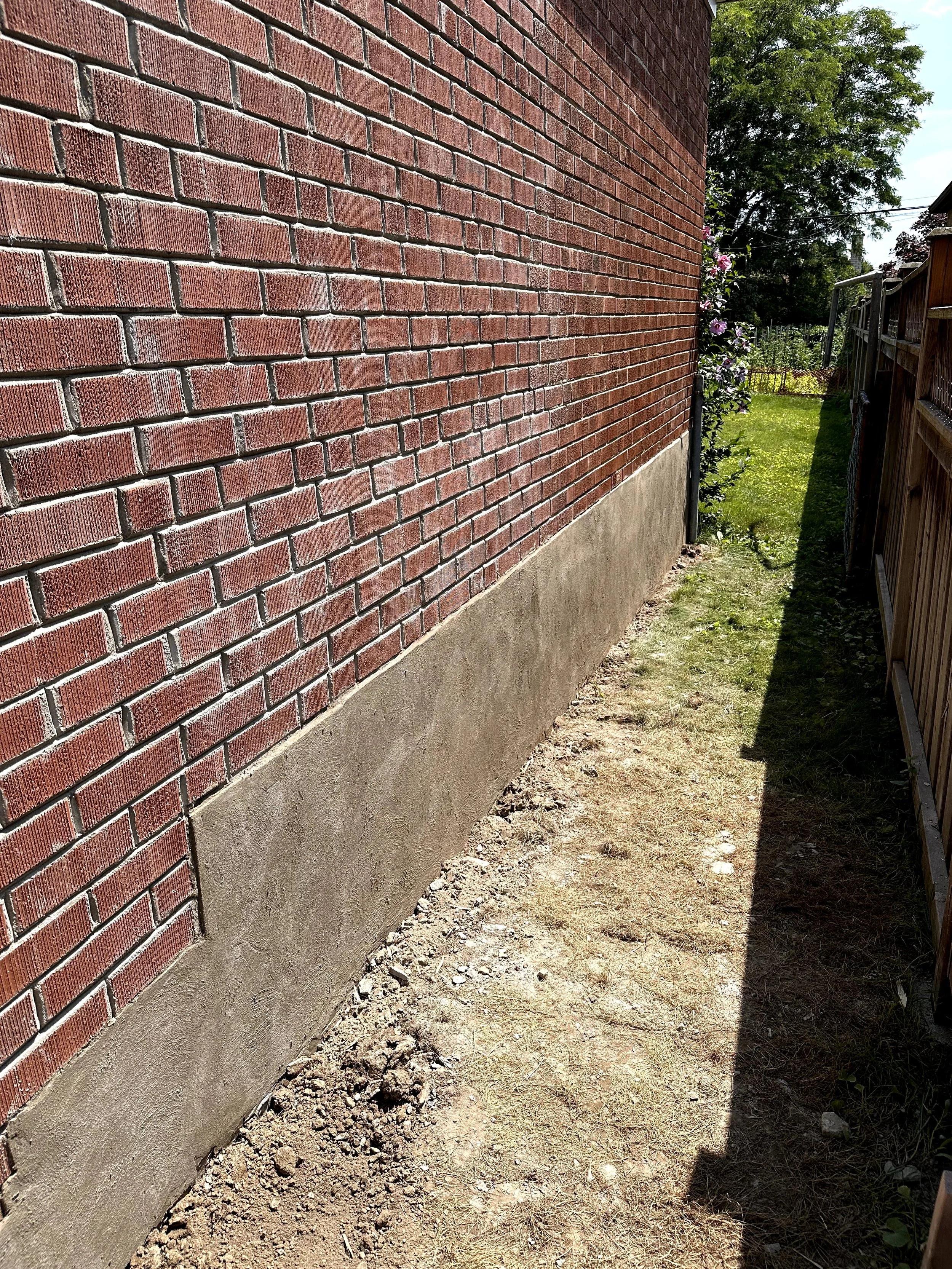 A brick wall and a small patch of dirt and grass beside it, with a wooden fence on the right and some plants and trees in the background.
