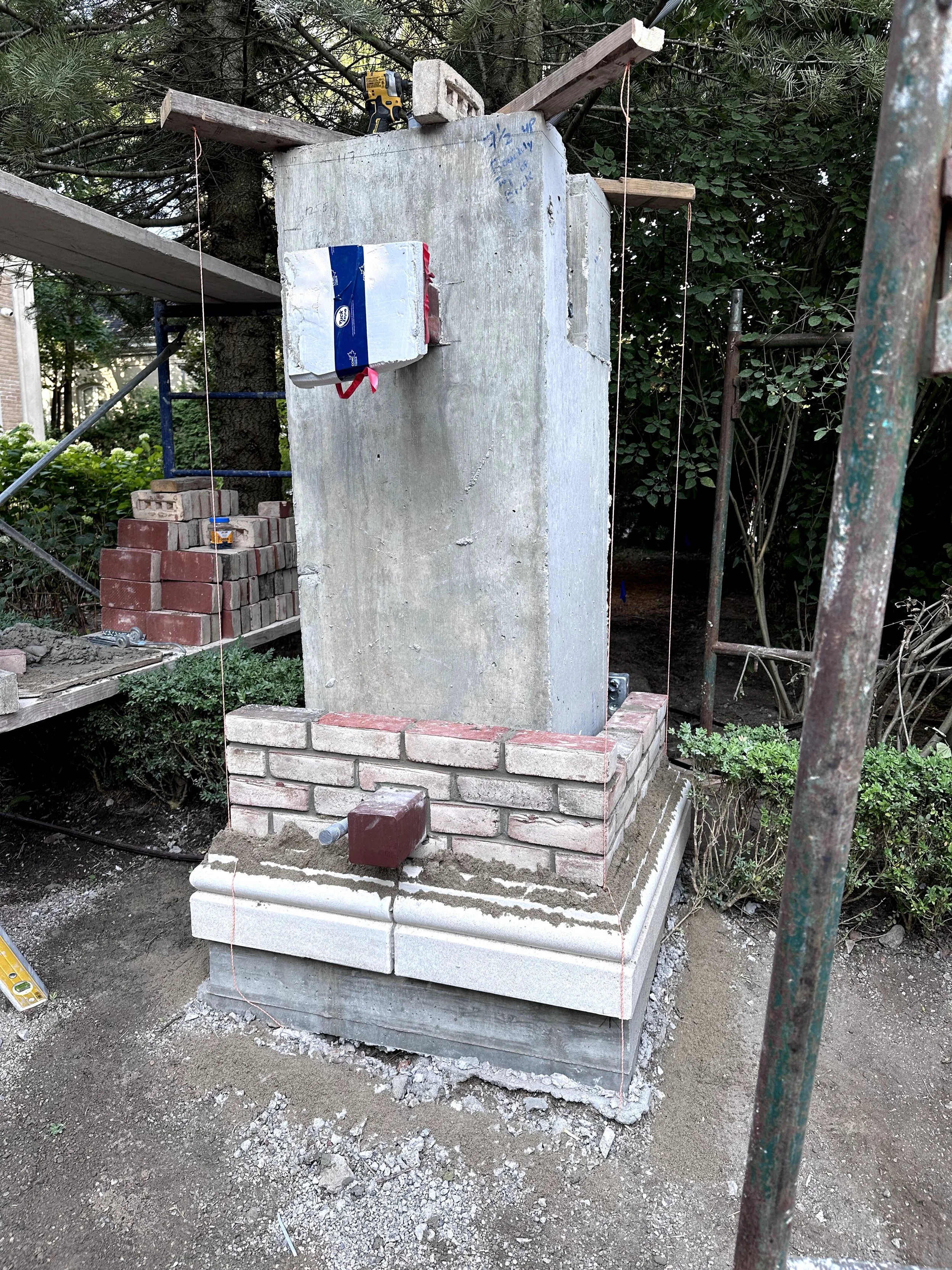 Construction site showing a large concrete pillar with wooden supports, bricks, and construction tools nearby, surrounded by trees and metal scaffolding.