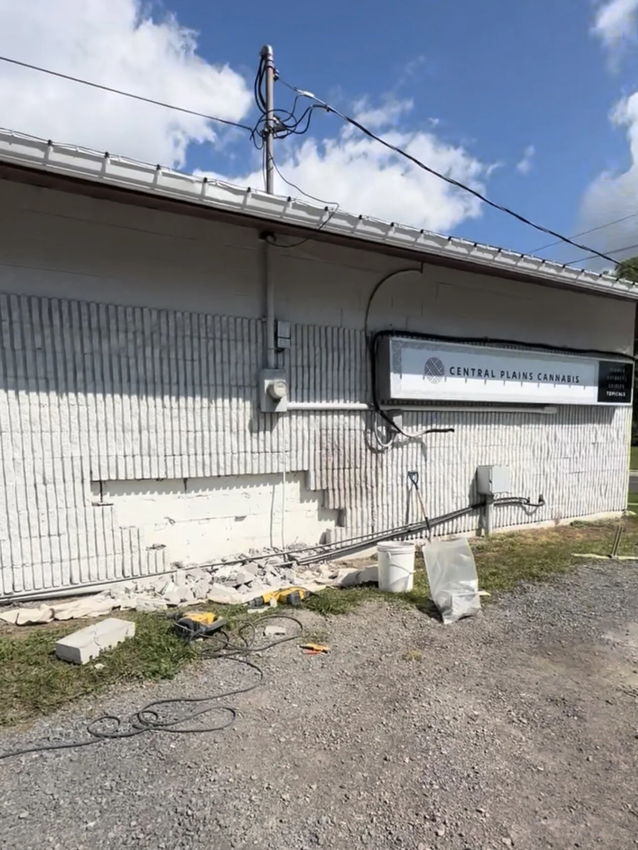 Outside of a building with a sign that reads 'Central Plains Cannabis.' Wiring and electrical boxes are mounted on the exterior wall, with some construction tools and debris on the ground.