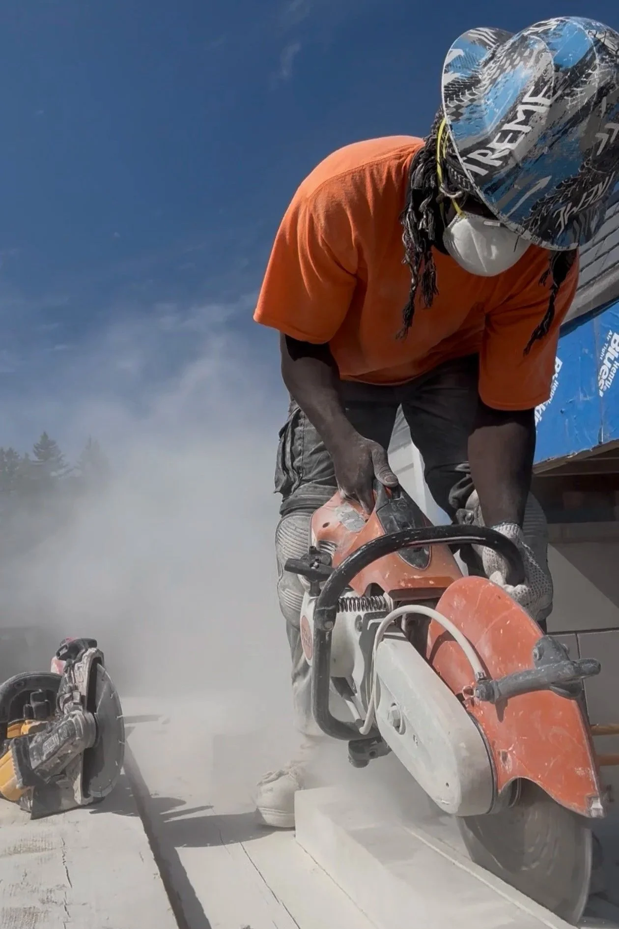 A person wearing an orange shirt, a helmet, a face mask, and gloves is operating a saw to cut a piece of wood. Dust is flying around due to the cutting process, and a truck with blue markings is in the background.