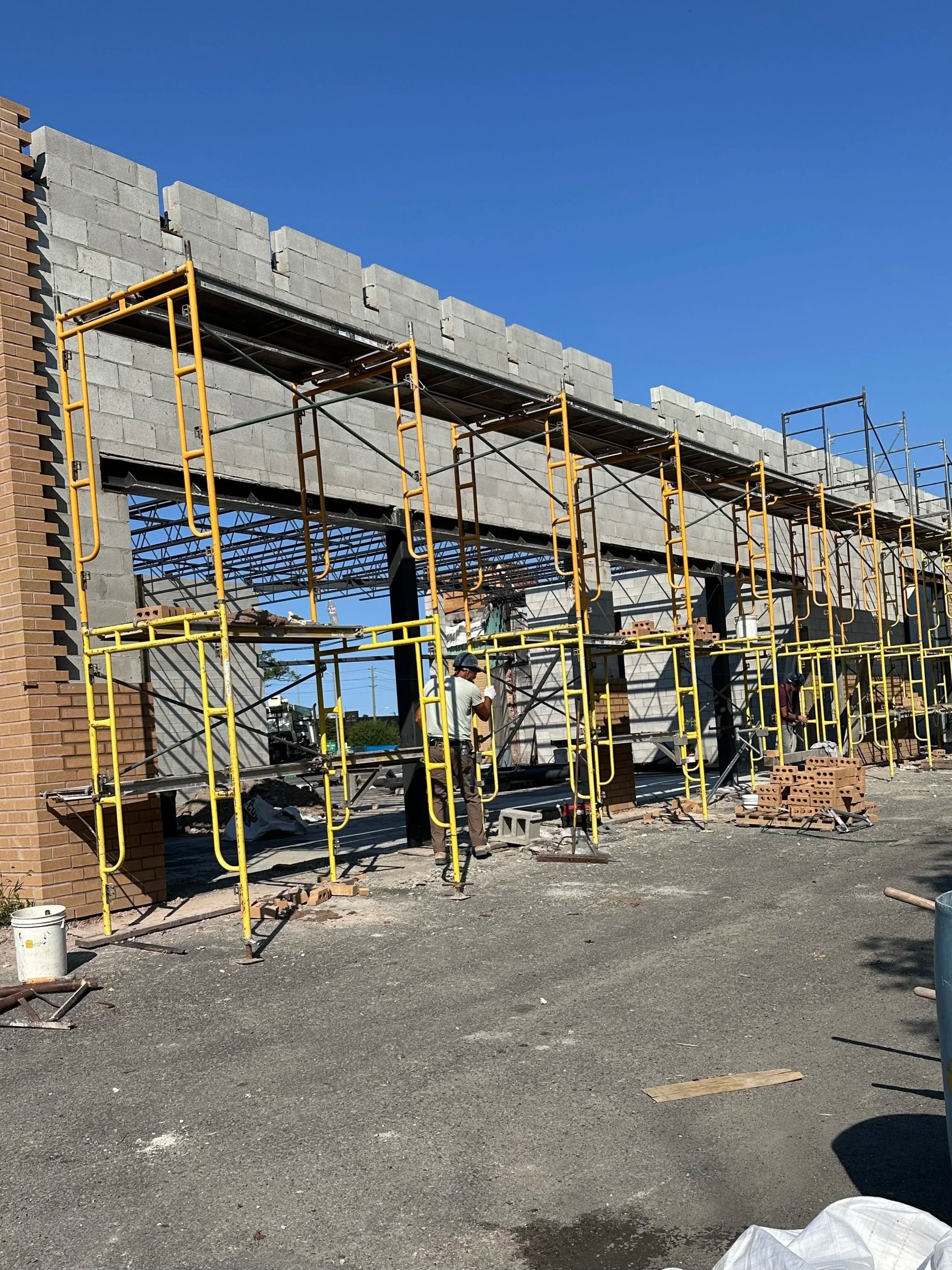 Construction site with scaffolding and workers building a concrete wall under a clear blue sky.