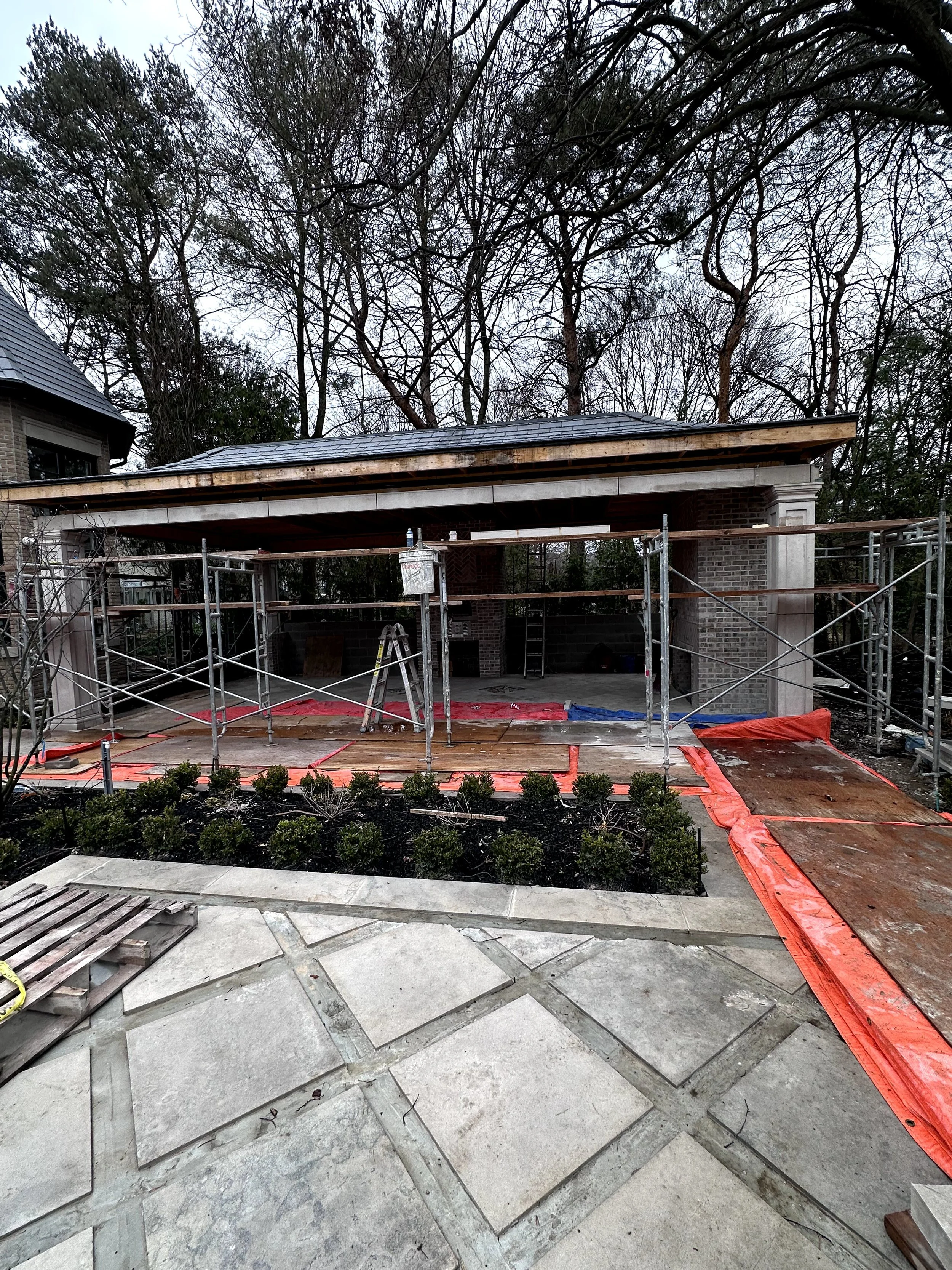 Construction site with scaffolding, a partially built brick structure, a roof extension, and a landscaped area with shrubs in the foreground, and trees with bare branches in the background.