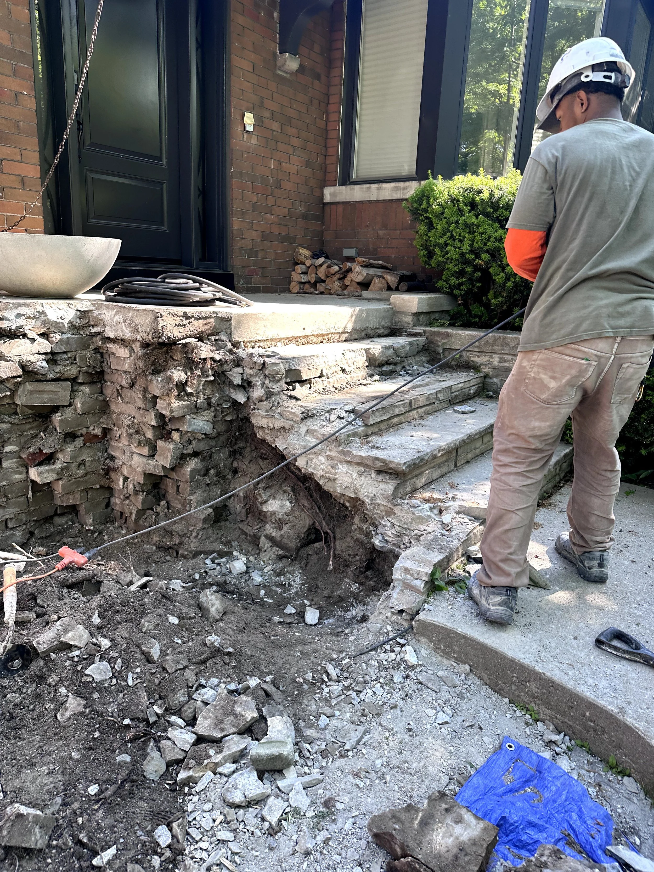A construction worker wearing a safety helmet and work gloves is demolishing or repairing concrete stairs outside a brick house. The stairs are partially broken, and debris and rocks are scattered around. A pile of firewood and a shrub are visible ne
