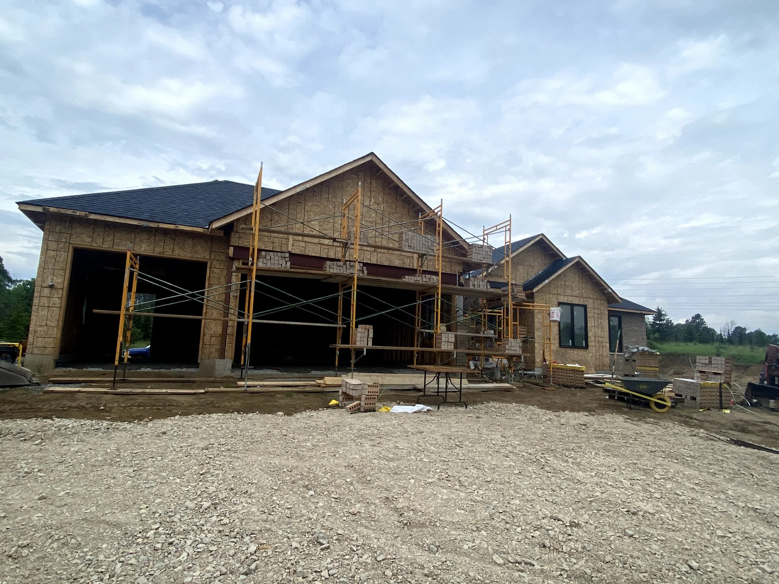 A house under construction with scaffolding, brick walls, a pitched roof with dark shingles, large windows, and construction materials around it, set against a cloudy sky.