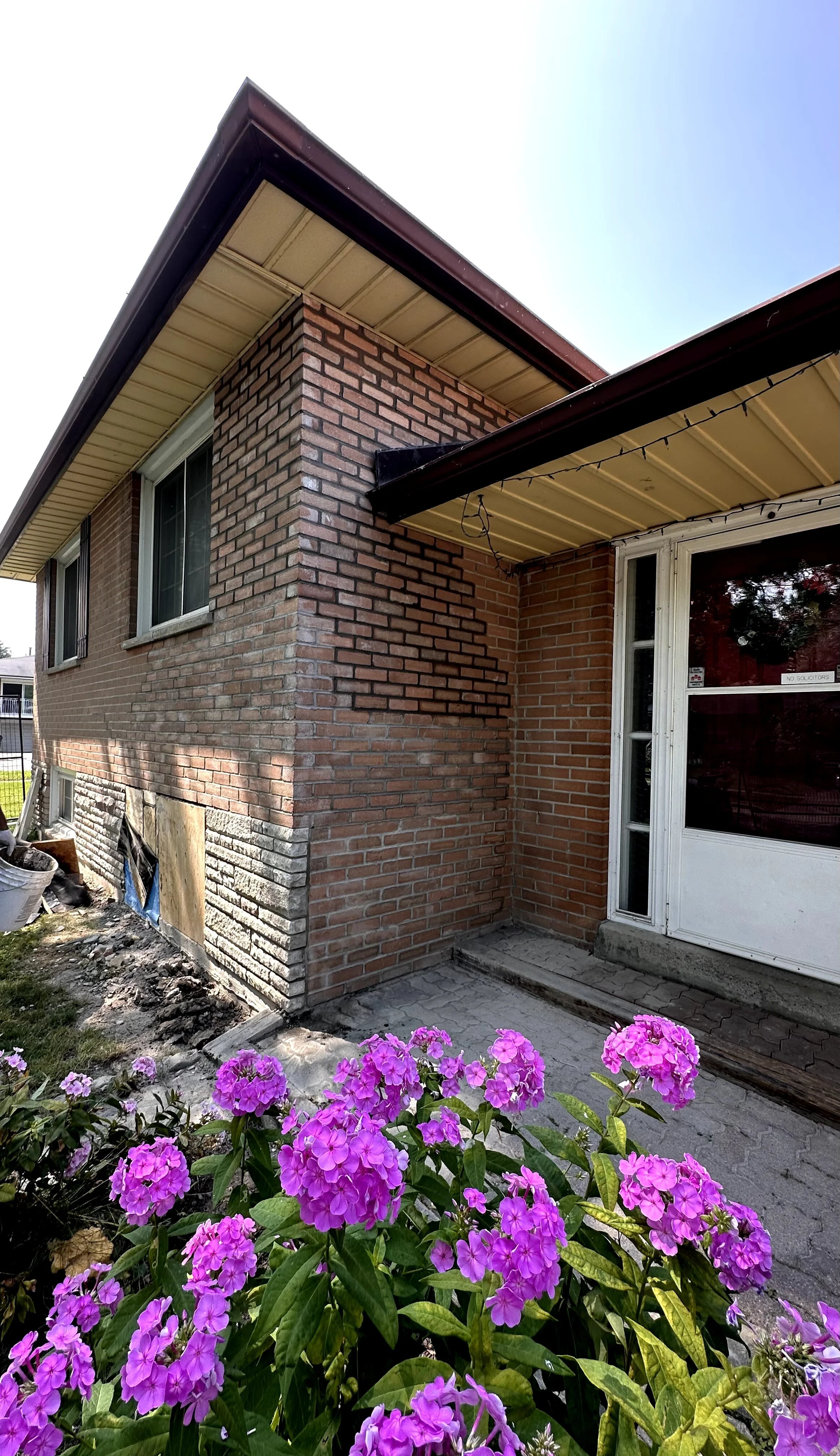 The image shows the exterior of a brick house with a small flower garden of pink and purple flowers in the foreground. The house has a window and a sliding glass door, with a section of the house under renovation or construction with some constructio