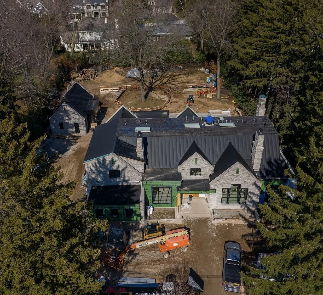 Aerial view of a large house under construction, surrounded by trees, with construction equipment and materials scattered around the yard.