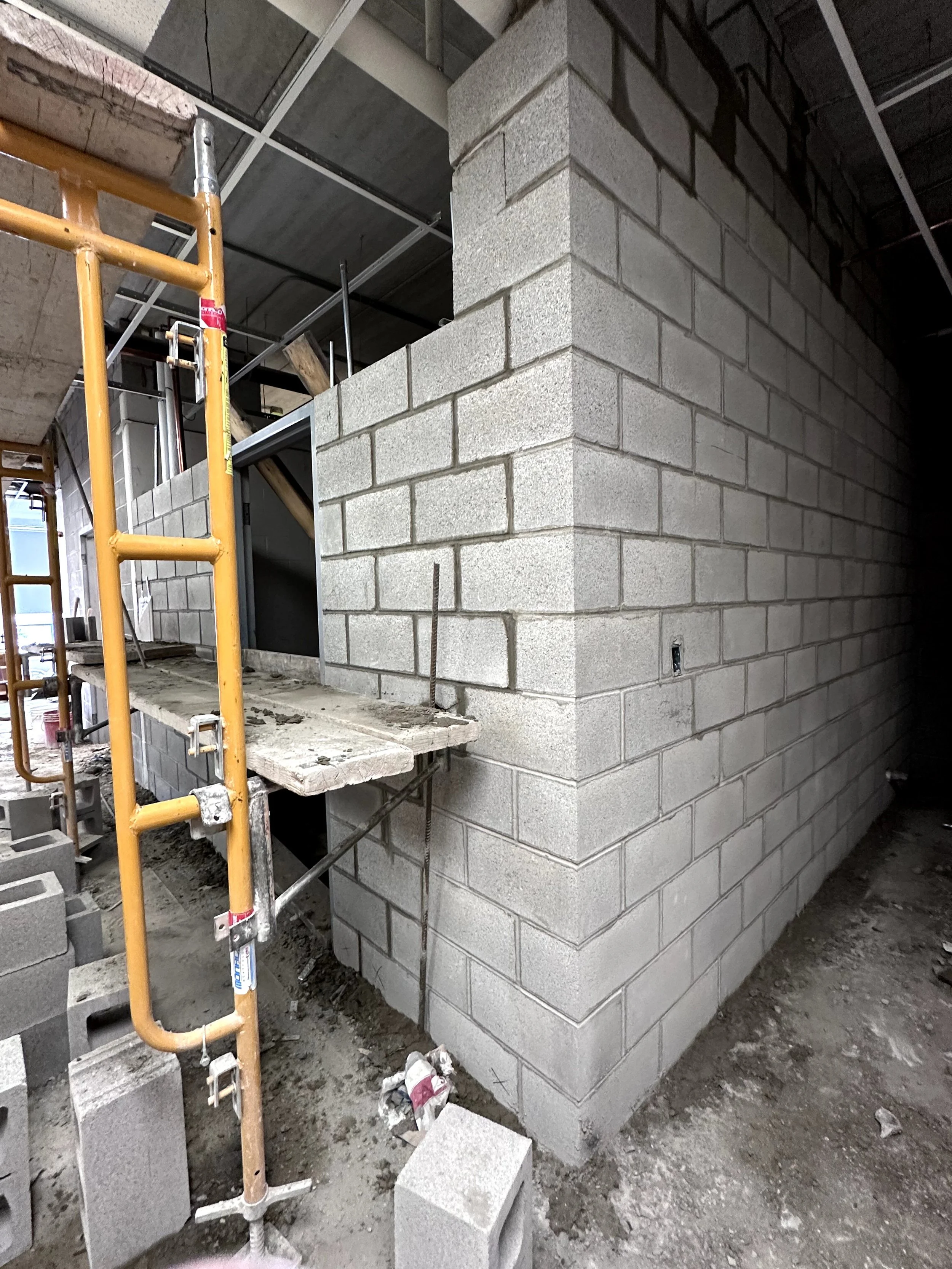 Construction site showing a partially built concrete block wall, scaffolding, and construction materials on the ground.