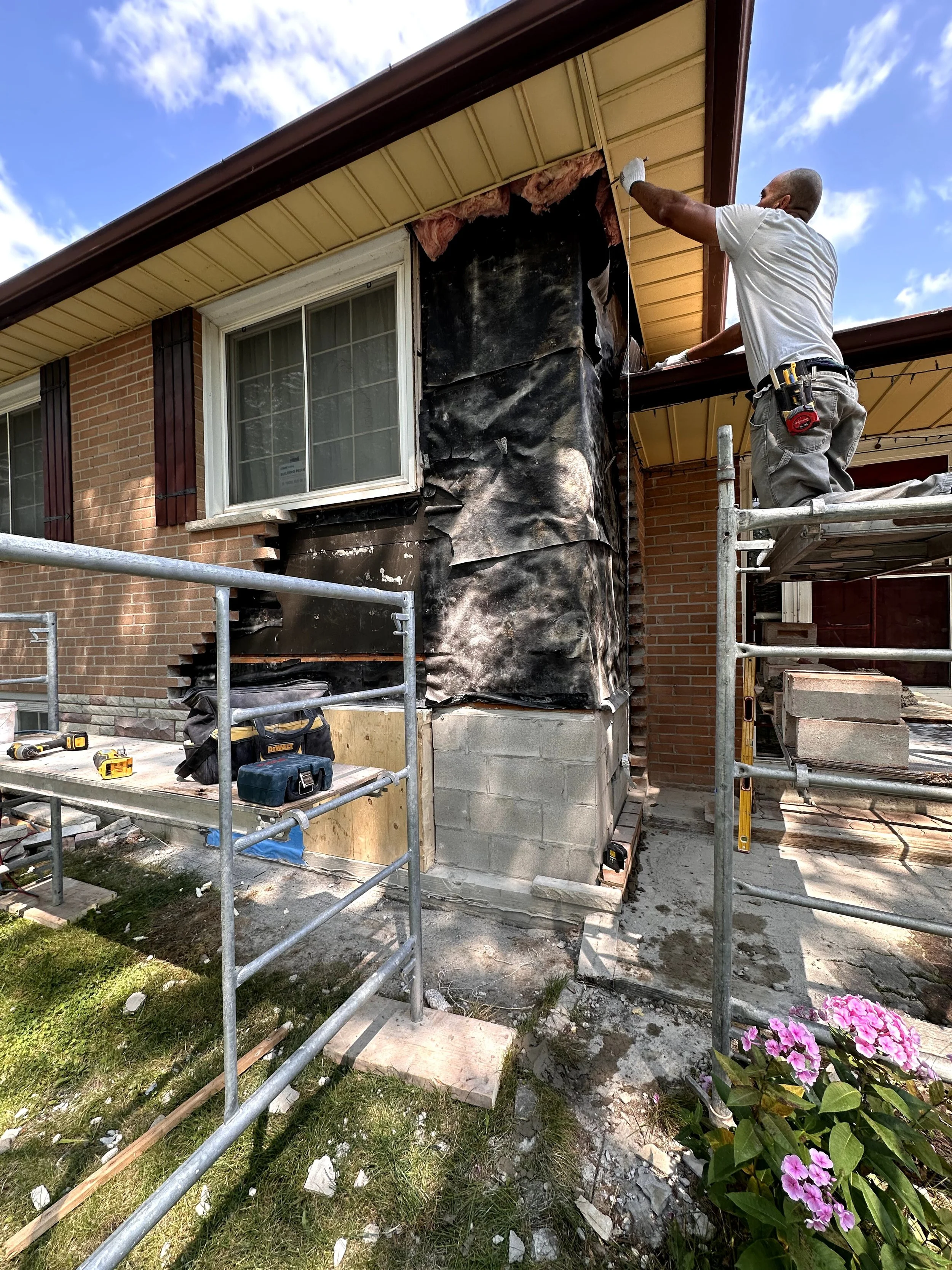 A construction worker standing on scaffolding repairing or inspecting the outside of a house with brick walls. The worker is bald, wearing a gray shirt and gray pants, and holding a tool. The house has a bay window, a beige roof with brown trim, and 