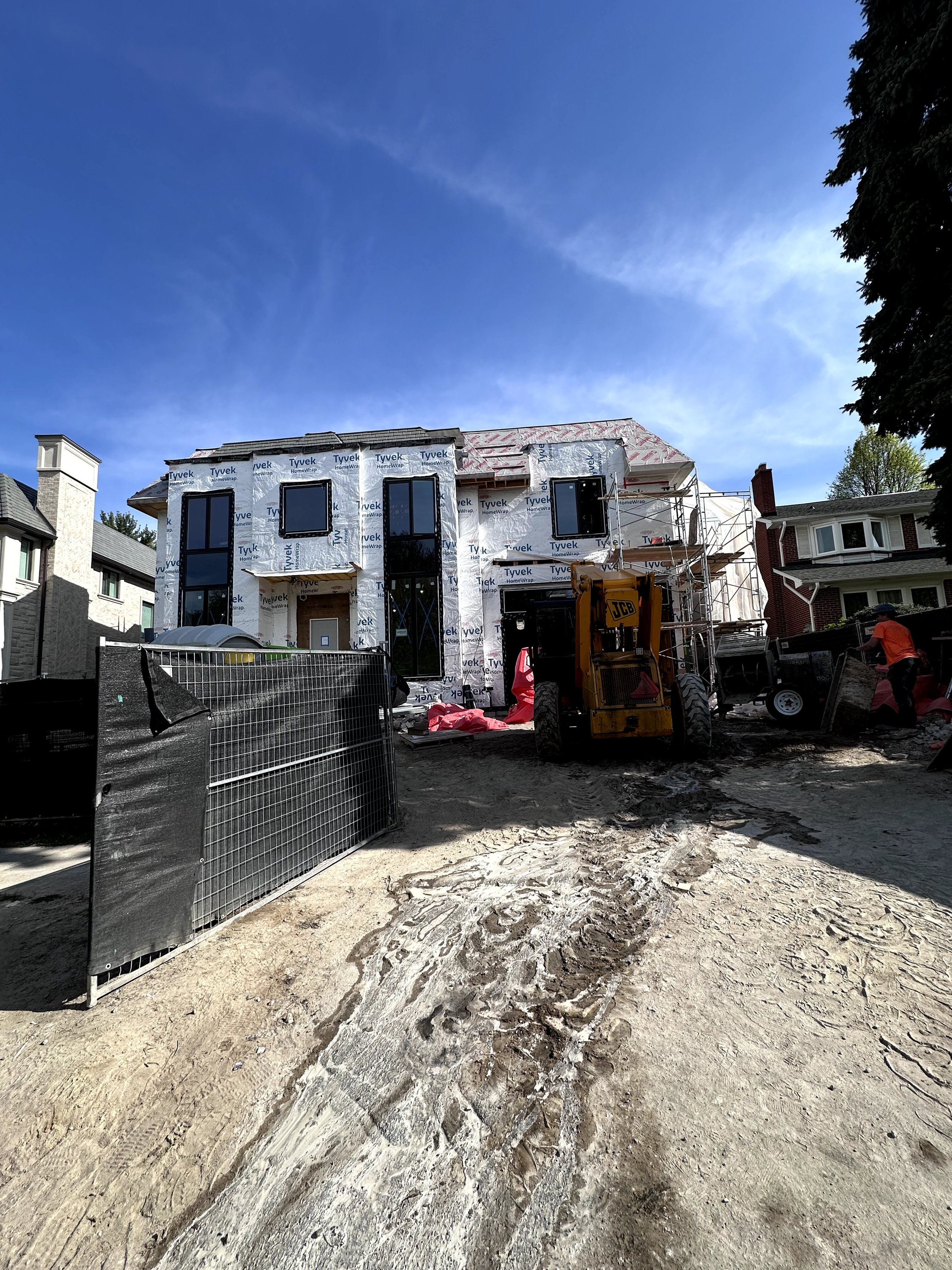 Under construction house with scaffolding and construction materials in front, surrounded by neighboring houses and a clear blue sky.