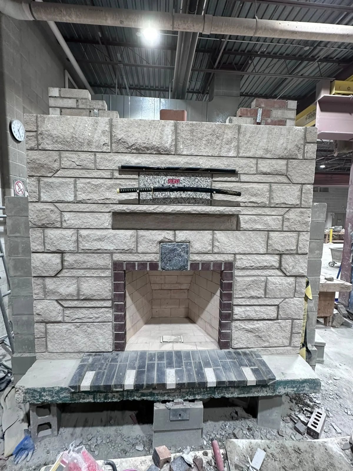 A partially constructed brick fireplace with a large stone mantel and a built-in chimney space, inside a workshop or construction site.