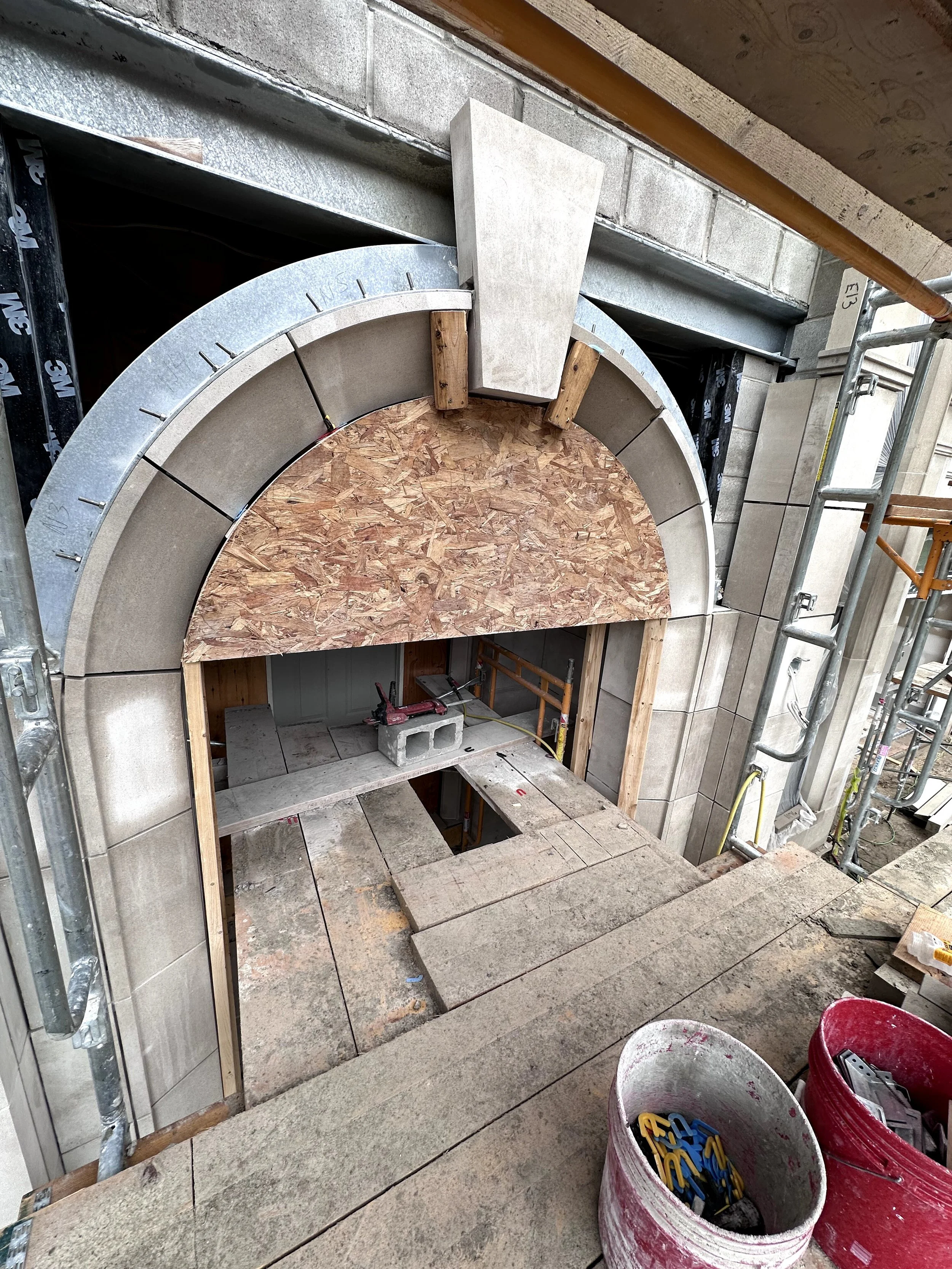 Construction site with a doorway, partially built arch with plywood and concrete blocks, scaffolding, and various construction tools and materials.