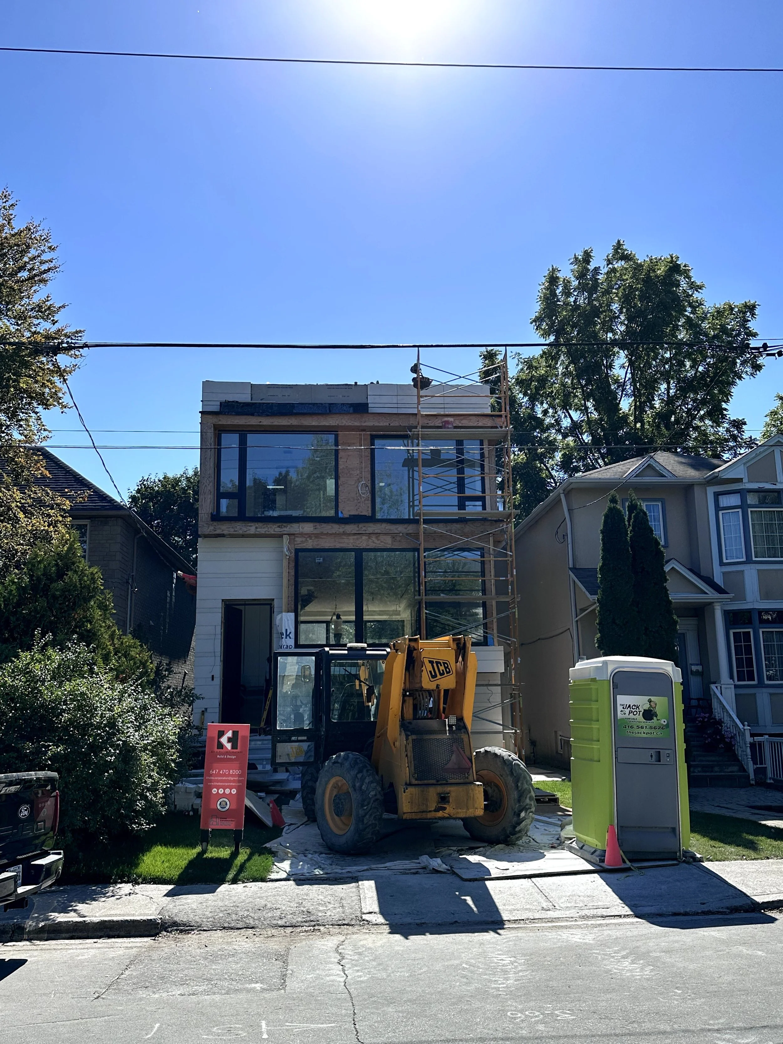 A house under construction with scaffolding on the front, a yellow JCB construction vehicle, a portable toilet, and construction signs in the driveway on a sunny day.