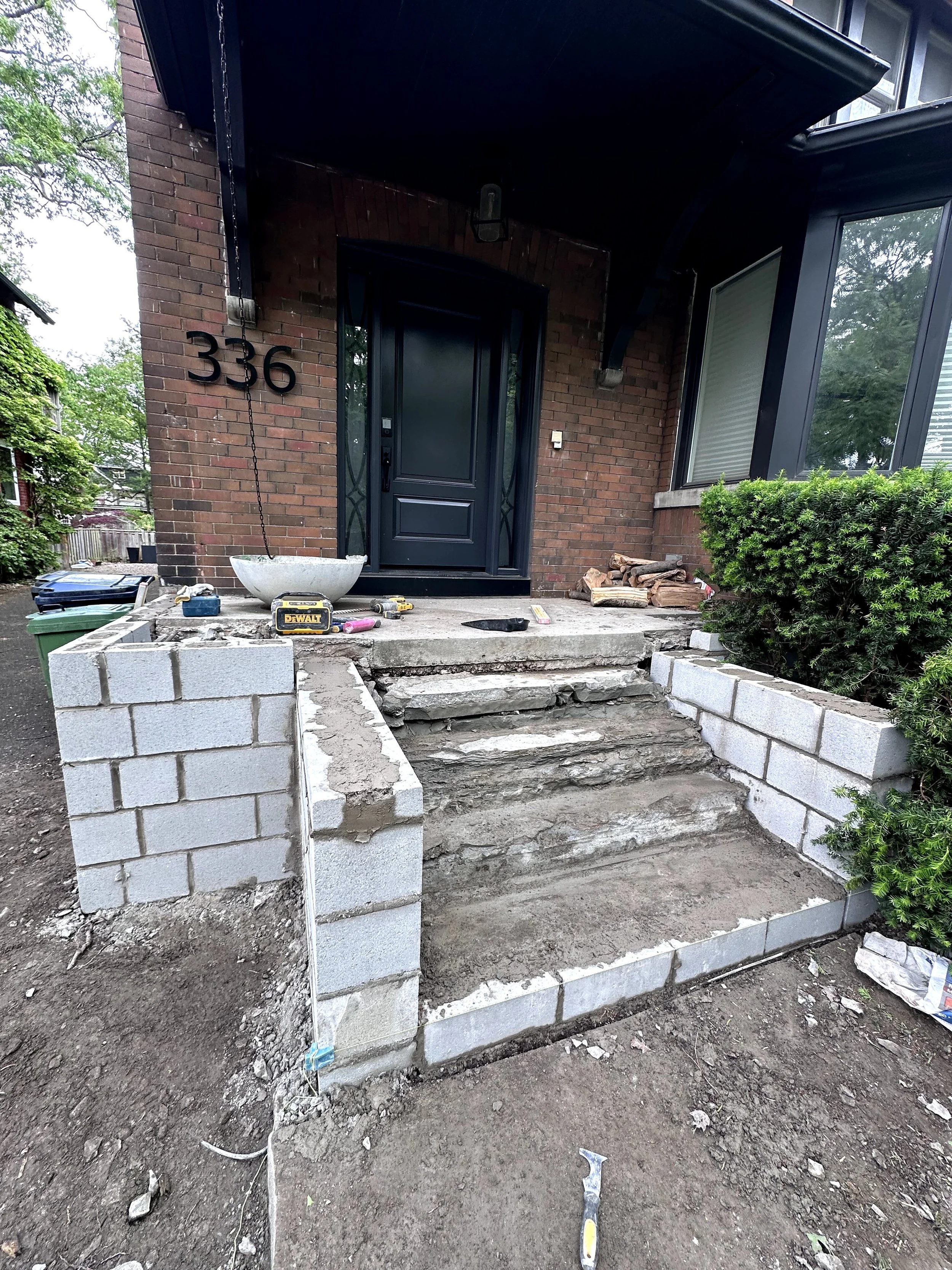Front porch under construction with concrete block walls, steps, construction tools, and a black front door on a brick house.