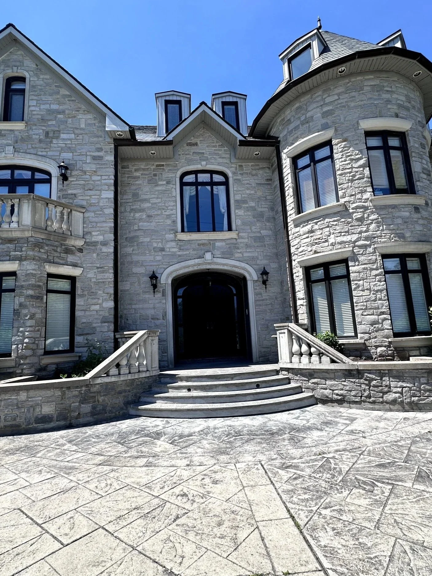 Front entrance of a large stone house with black-framed windows, a staircase leading up to the door, and a paved walkway in front.
