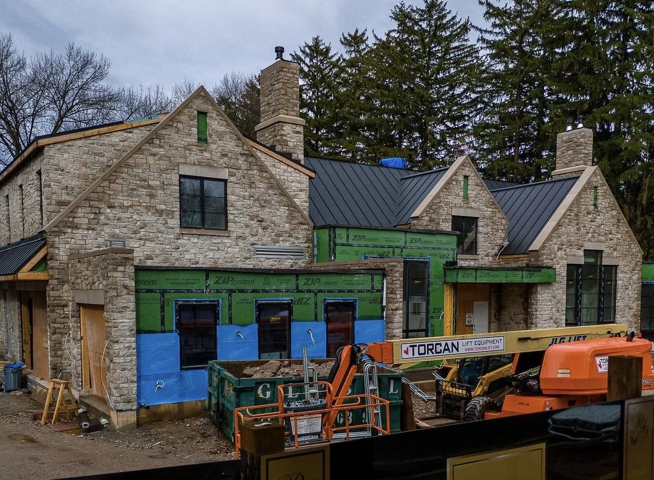 A house under construction with stone walls, green insulation panels, and black windows, surrounded by construction equipment and trees in the background.