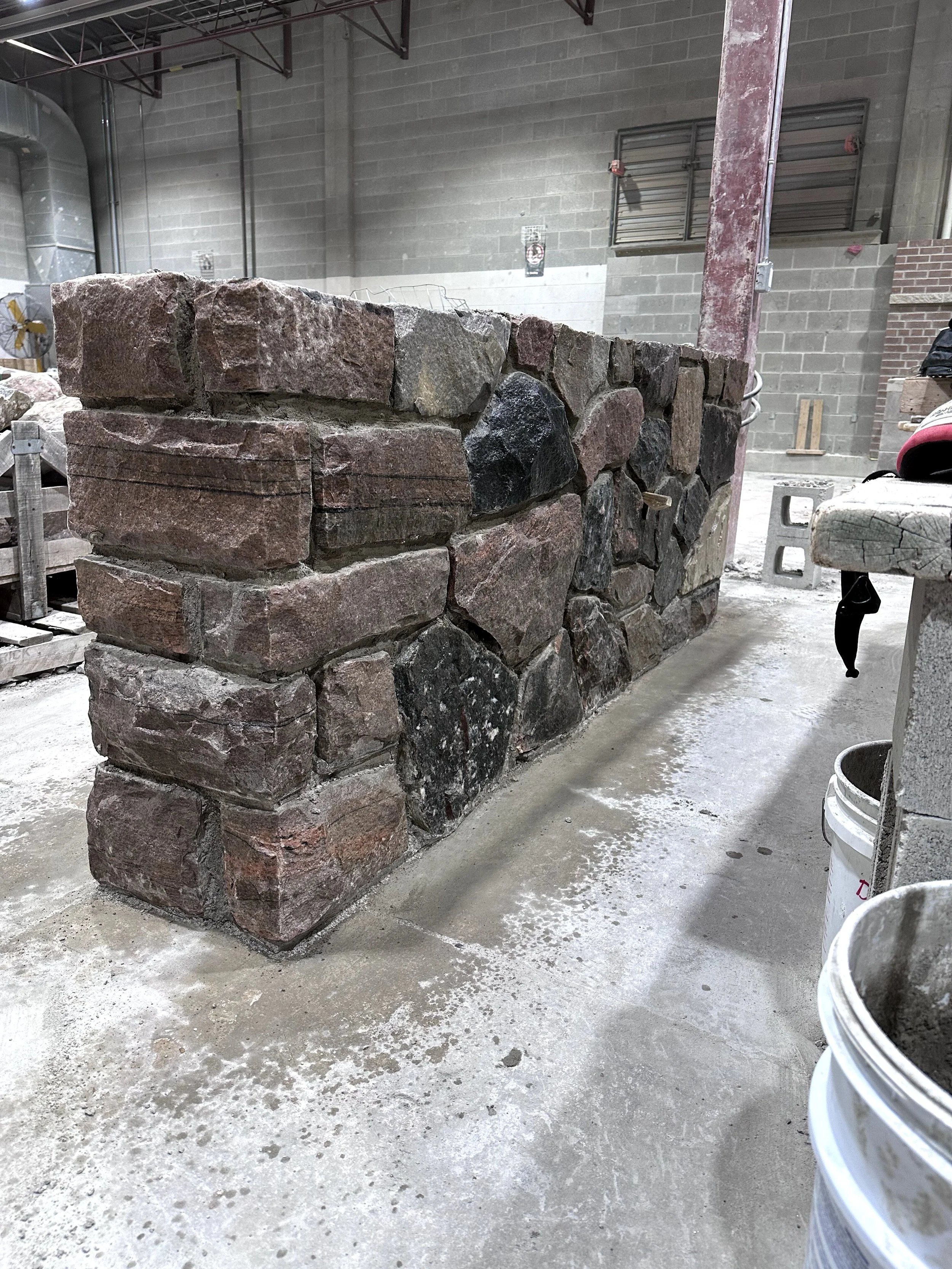A partially built brick and stone wall inside a construction warehouse. The wall is made of large, irregular stones and red bricks, with construction supplies and tools visible around.