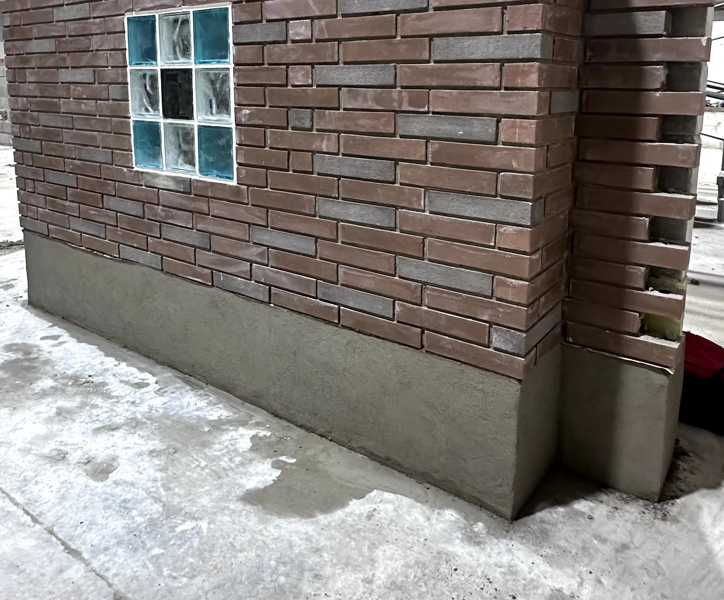 A close-up of a brick corner of a building with glass block window. The ground is icy and covered with snow.