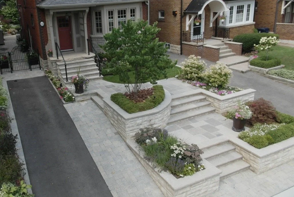 Front yard with a brick house, landscaped garden beds with shrubs and flowers, stairs, and a paved walkway.