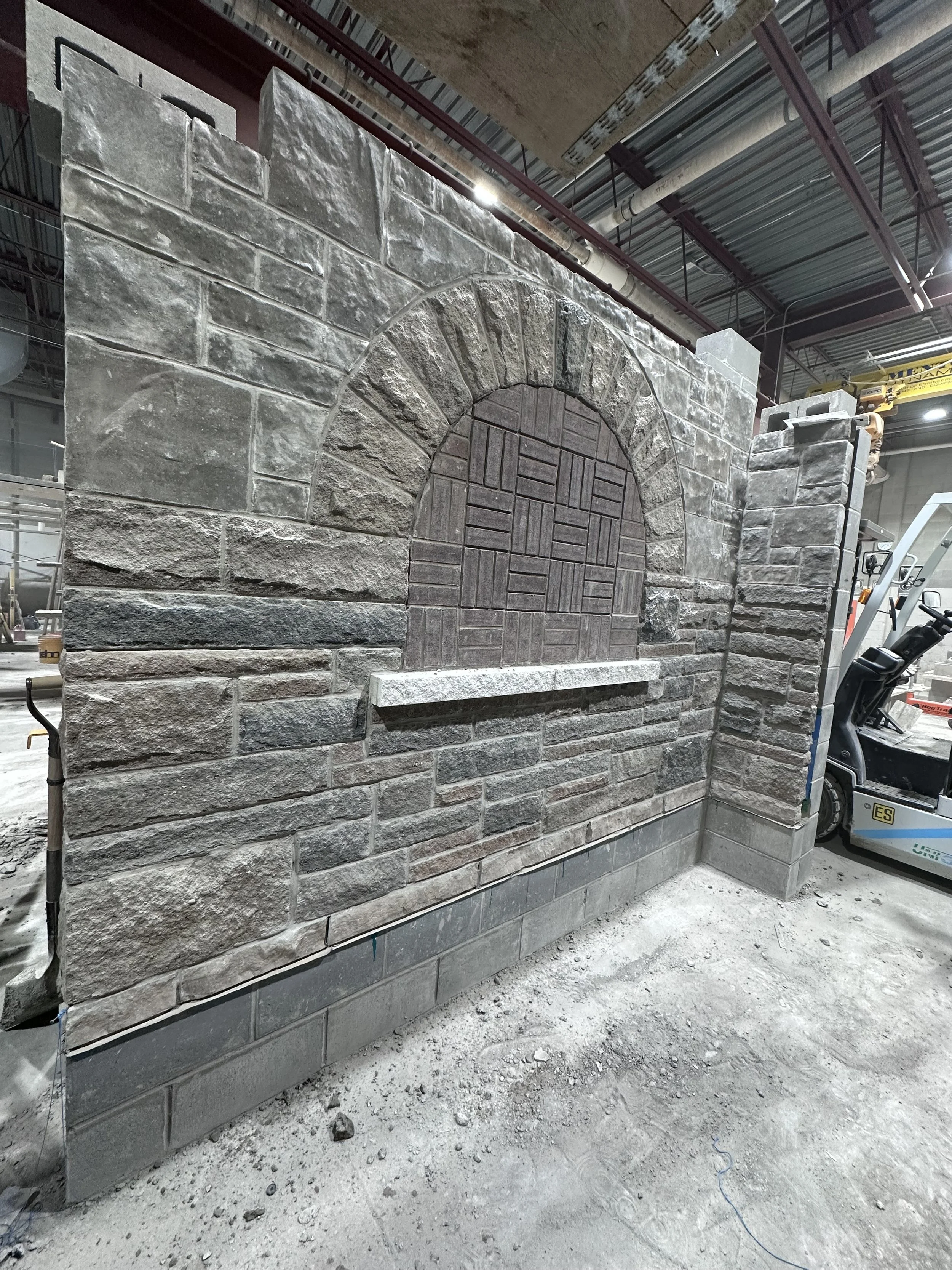 A partially built indoor stone wall with a decorative arch and a ledge, under construction in an industrial setting, with construction equipment and pipes visible overhead.
