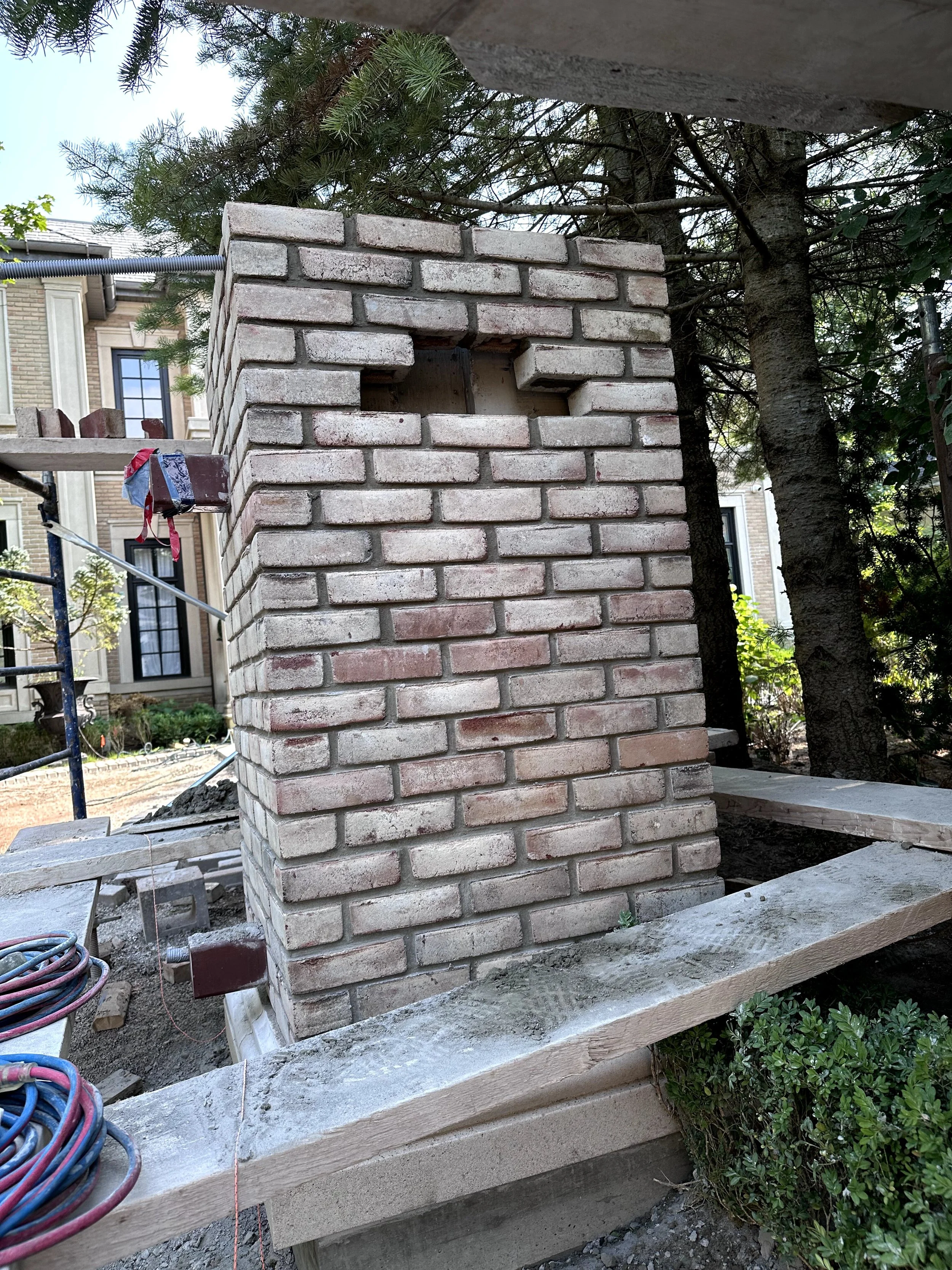 Close-up view of a brick chimney under construction, with bricks stacked and some construction materials and tools nearby, in an outdoor setting next to trees and a residential building.