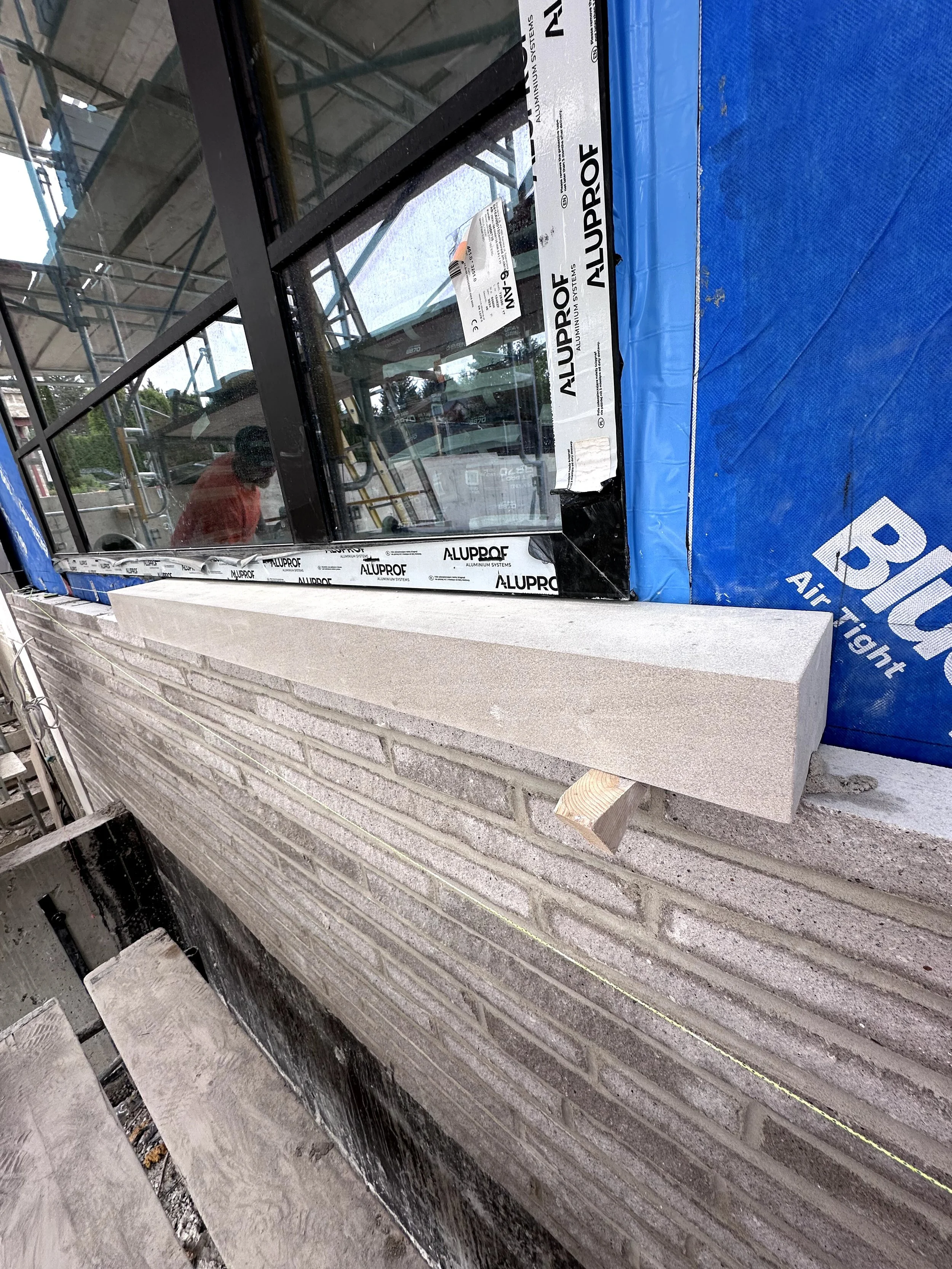 Construction site showing a window frame with aluminum systems, a person in an orange shirt, a brick wall, blue tarp, and construction tools on the ground.
