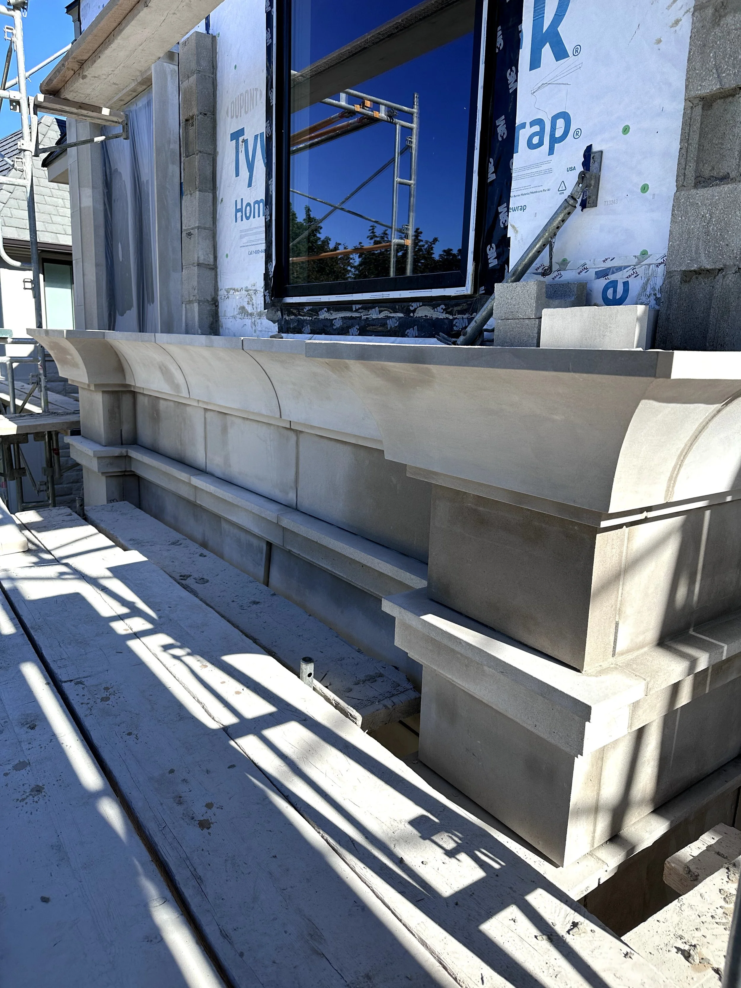 Construction site showing a house window area with scaffolding, and beige stone architectural elements being installed on the exterior wall.