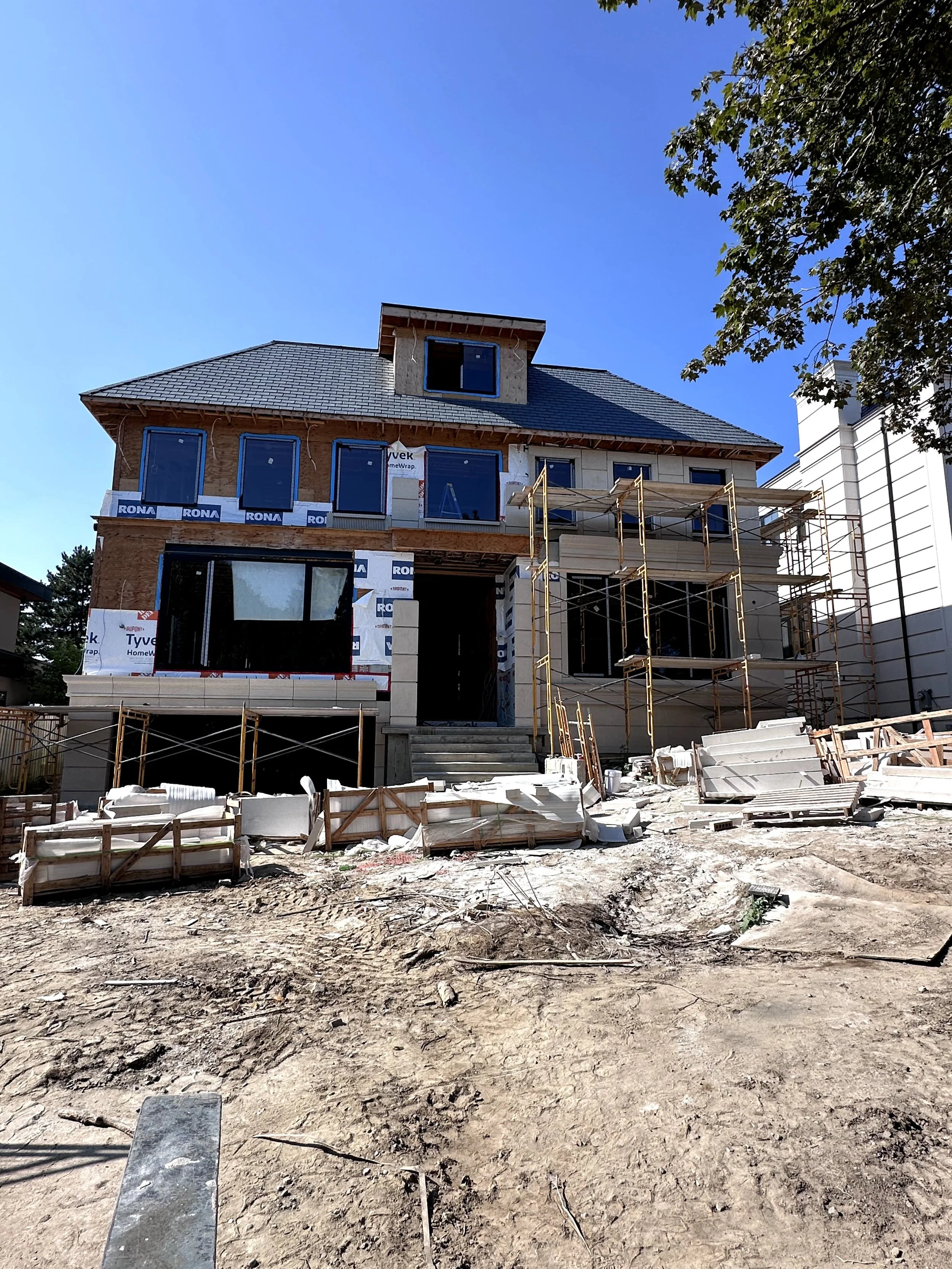 A house under construction with a partially completed exterior. Scaffolding surrounds the building, and construction materials are scattered on the ground. The sky is clear and blue, and a tree with green leaves is visible on the right side.