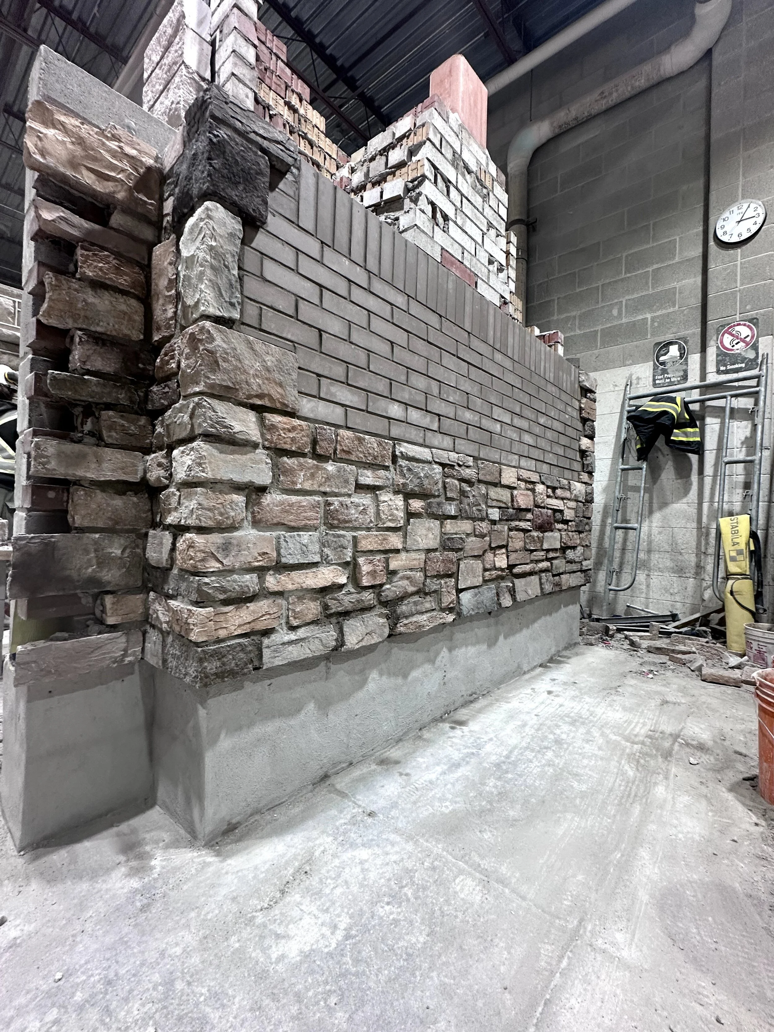 Construction site showing a partially built brick and stone wall with construction materials and safety equipment nearby.